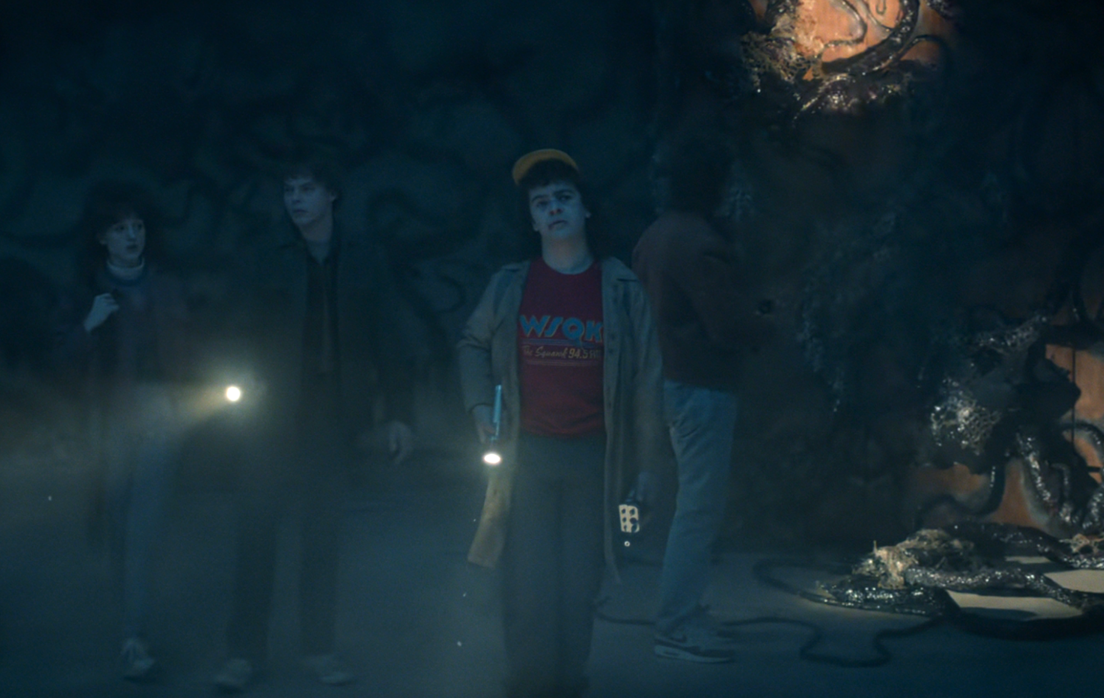 A group of four people explores a dark, ominous cave with flashlights, inspecting twisted roots on the walls