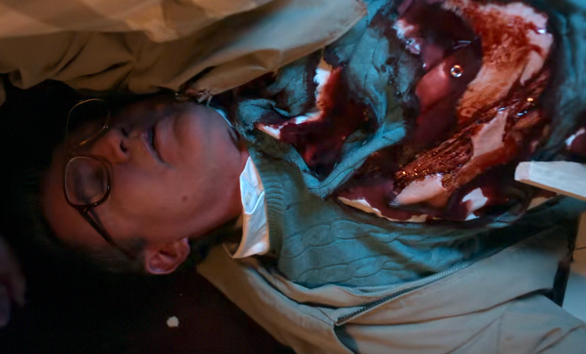 Person lying on the ground with glasses, wearing a cable-knit sweater and a trench coat, appearing unconscious with a simulated injury on their chest