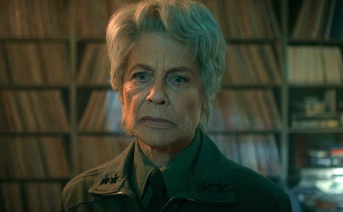 Person with short hair in a military-style jacket stands in front of a wall of records, looking serious
