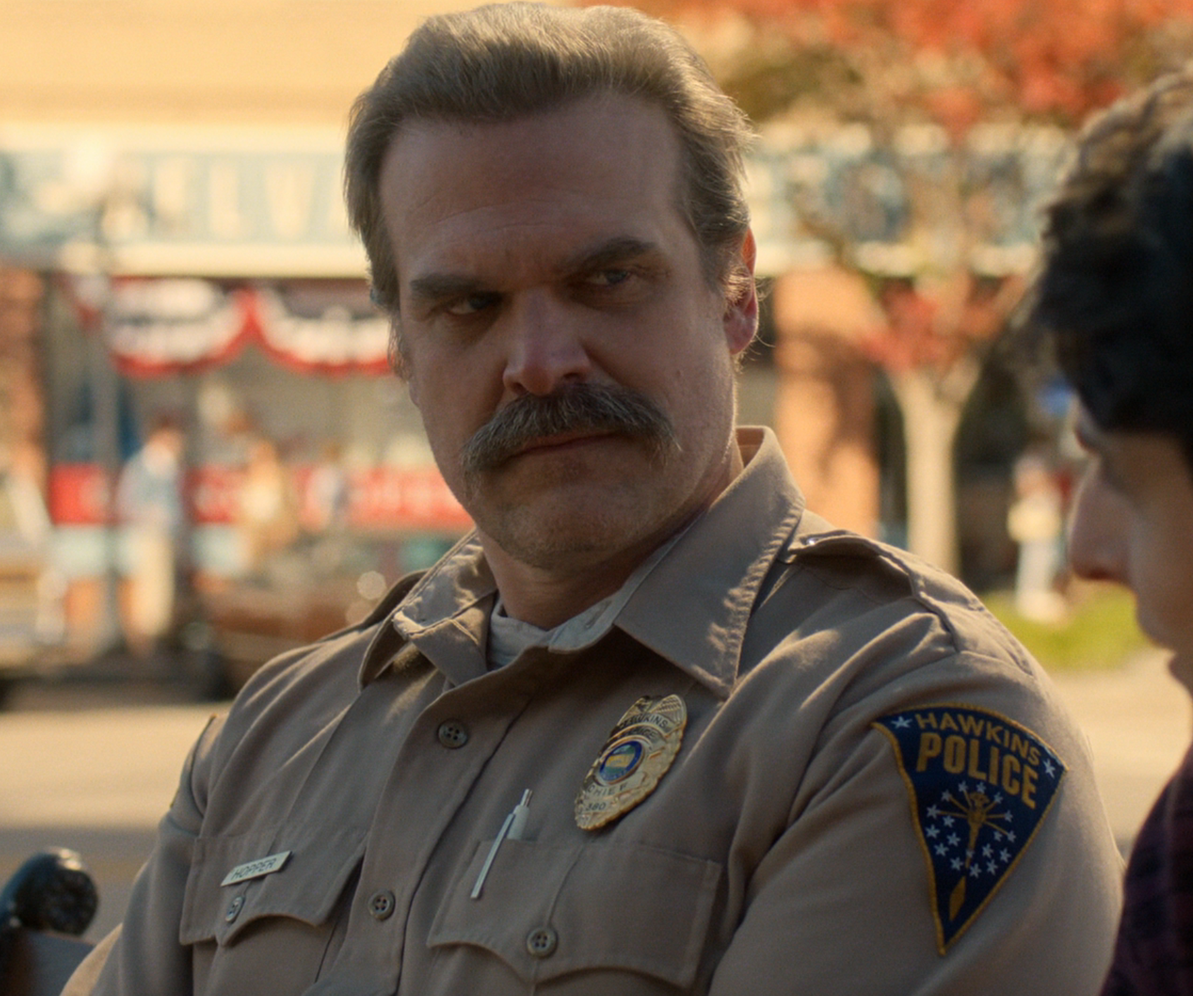 Man in police uniform with "Hawkins Police" badge talks to another person outside in a small town setting