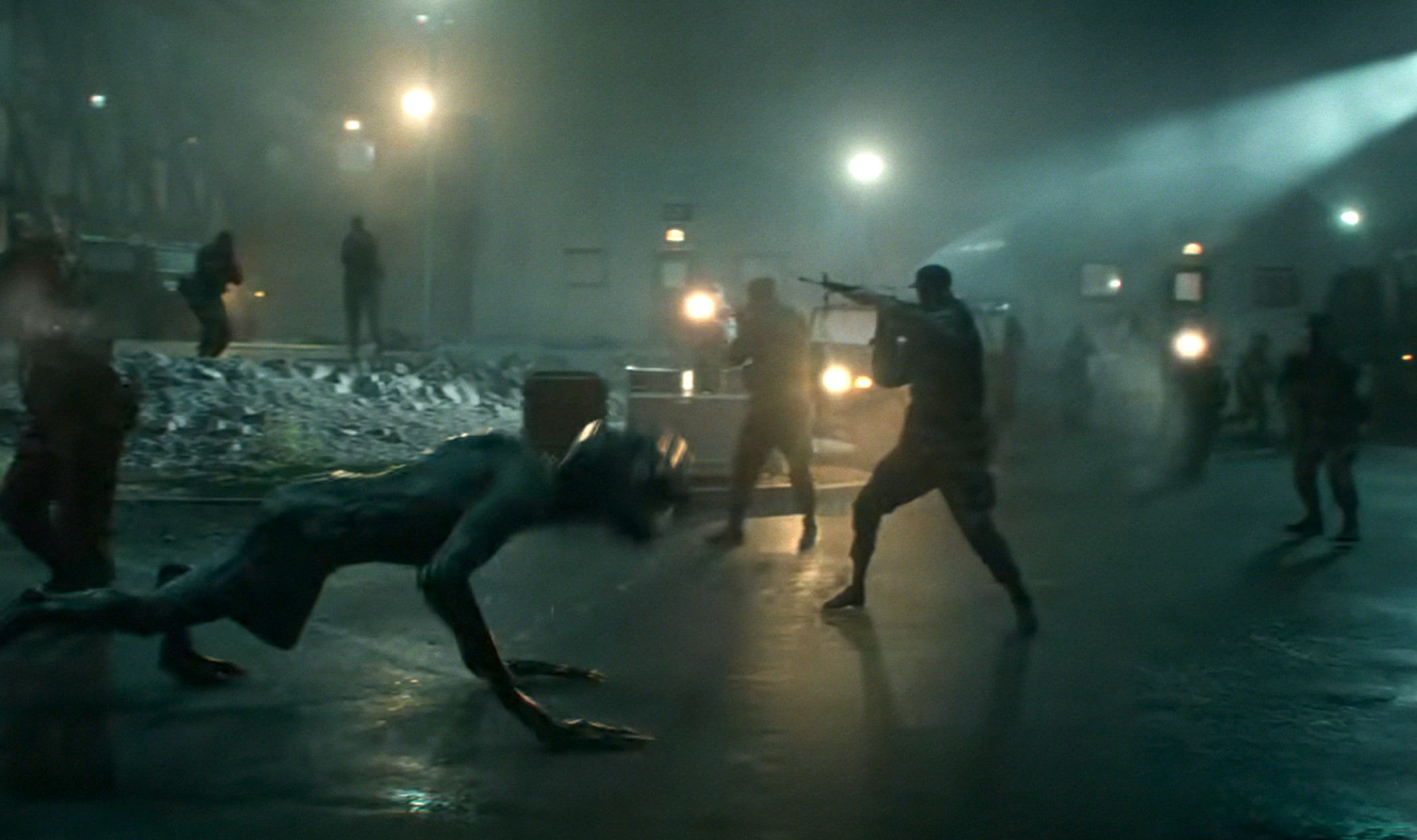 Action scene with people facing off against a crawling creature at night, with bright lights and snow in the background