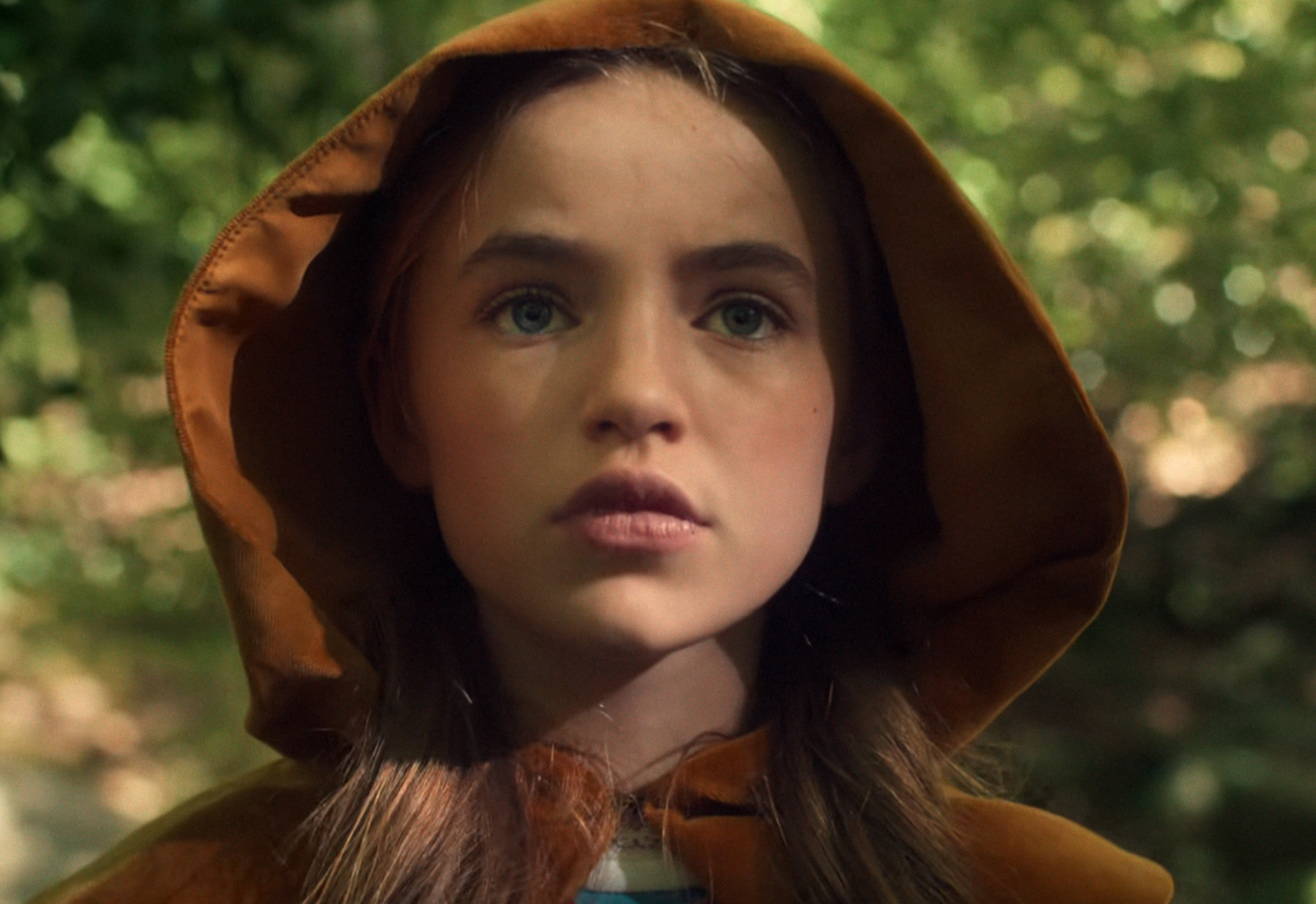 A child with long hair, wearing a hooded cloak, looks thoughtfully into the distance in a forest setting