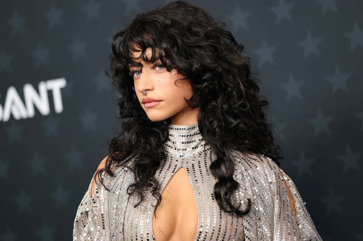 Person in a glamorous, intricately beaded gown with wavy, voluminous hair poses confidently at a formal event