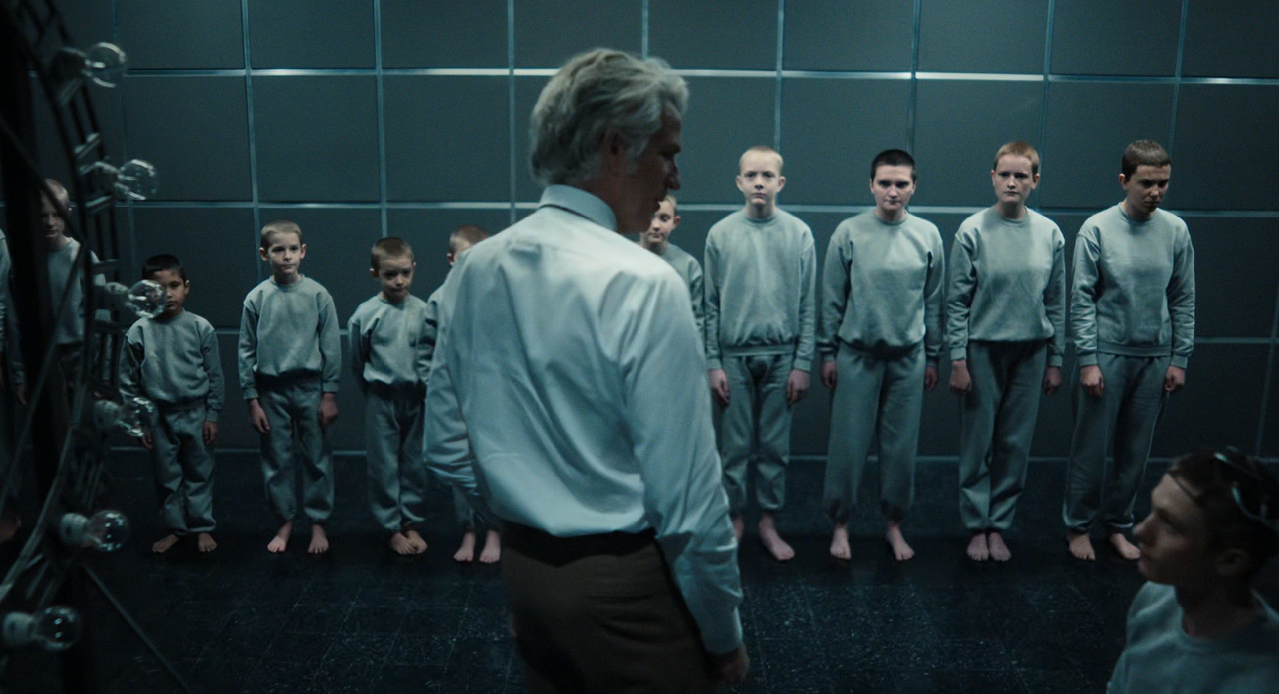 An older man in a dress shirt faces a line of children wearing identical gray outfits, standing barefoot against a tiled wall