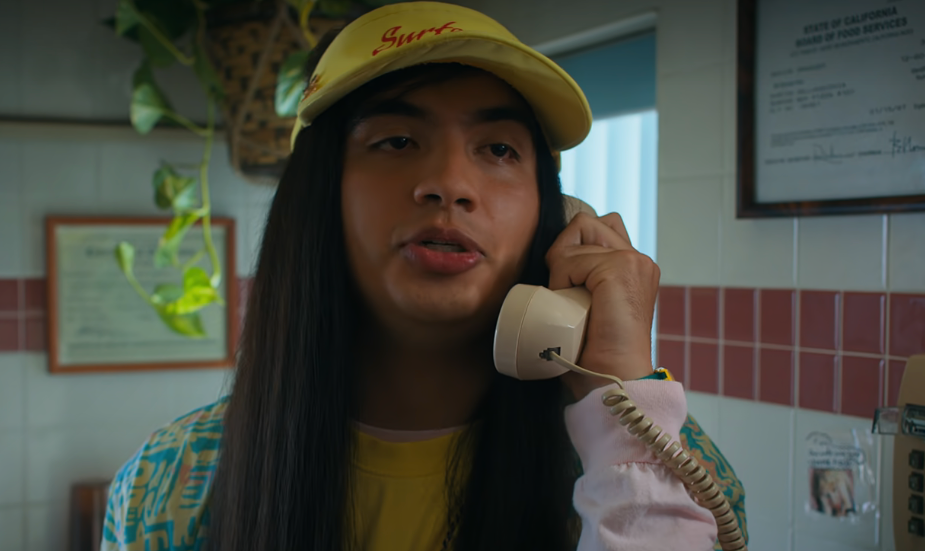 Person with long hair, wearing a yellow cap and casual patterned shirt, speaks on a corded phone in a room with framed certificates on the wall