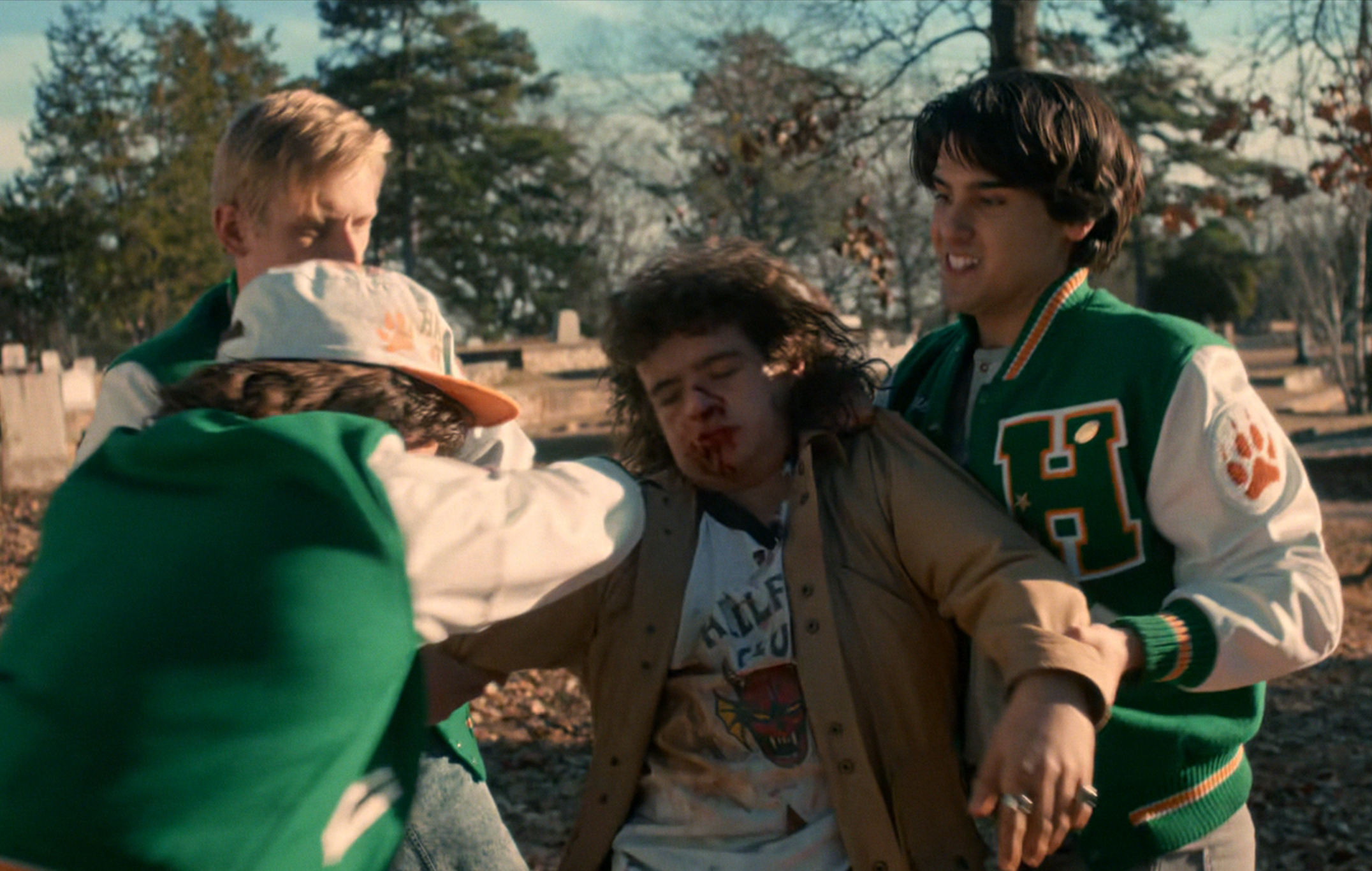 A group of teenagers wearing varsity jackets appear to be in a conflict, one seems hurt with a bloody nose, in an outdoor setting