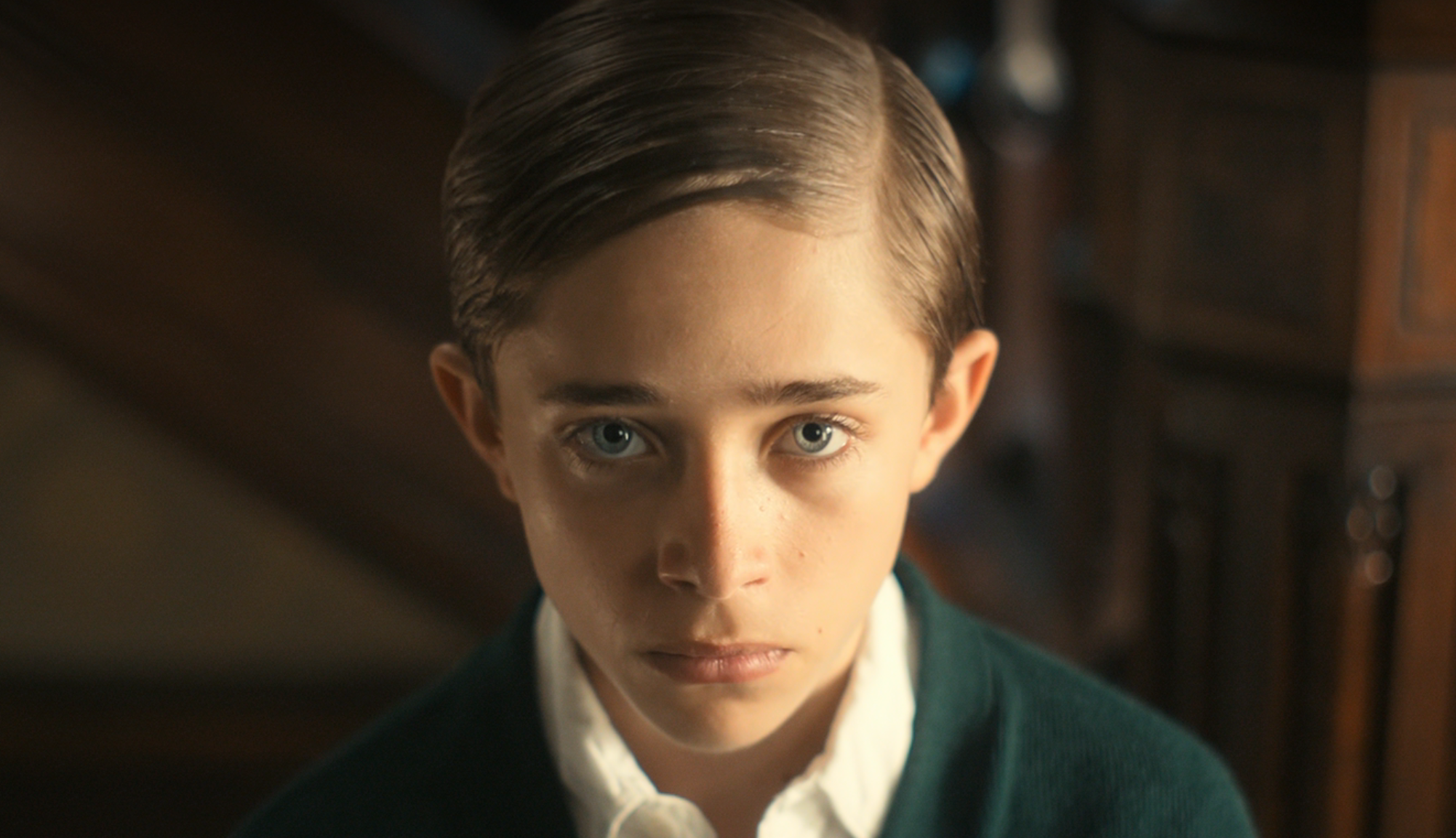 Young person with neatly combed hair looks directly at the camera, wearing a collared shirt and sweater, with a neutral expression