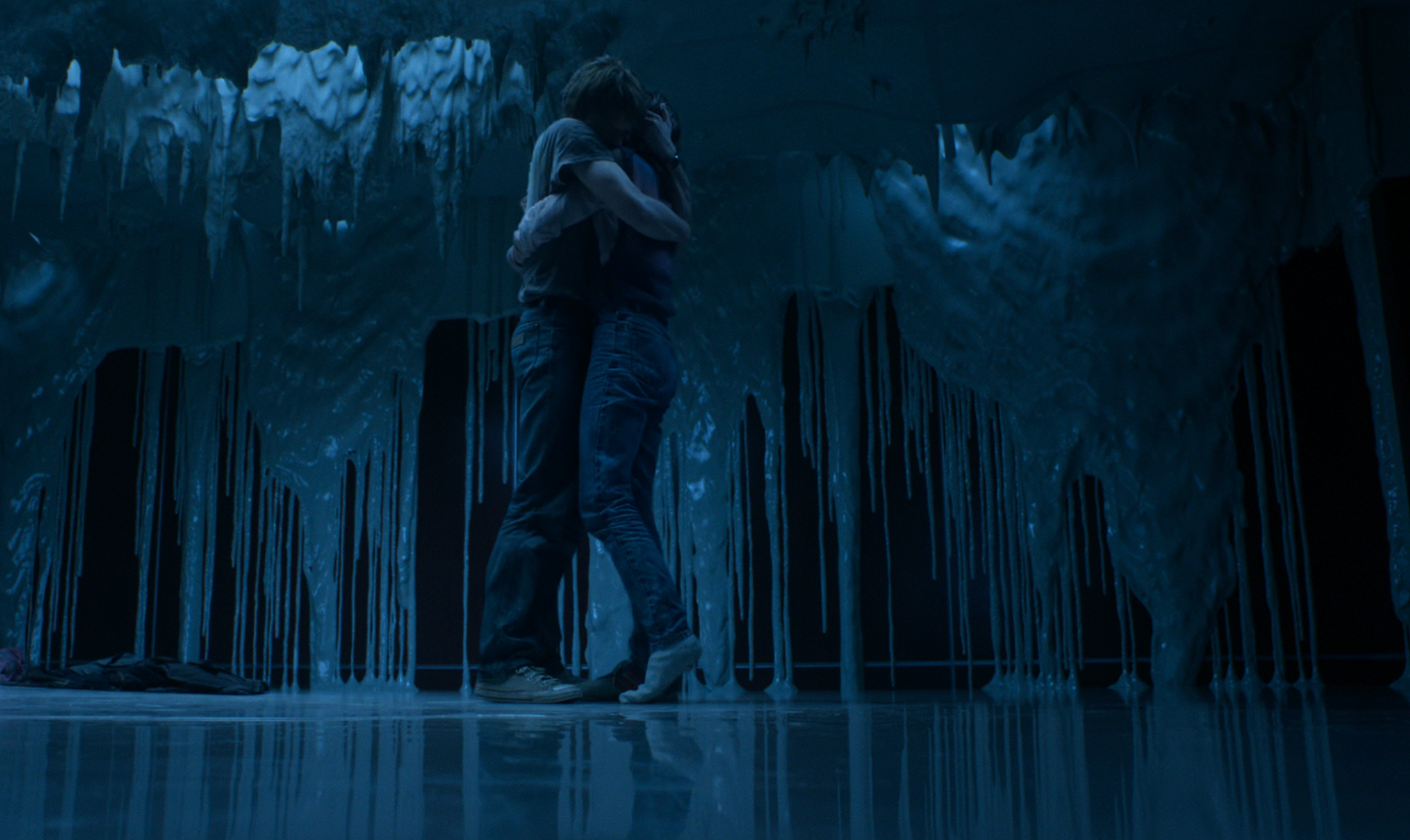 A couple embraces in the center of a room with cascading, icy-like structures from the ceiling giving a surreal atmosphere