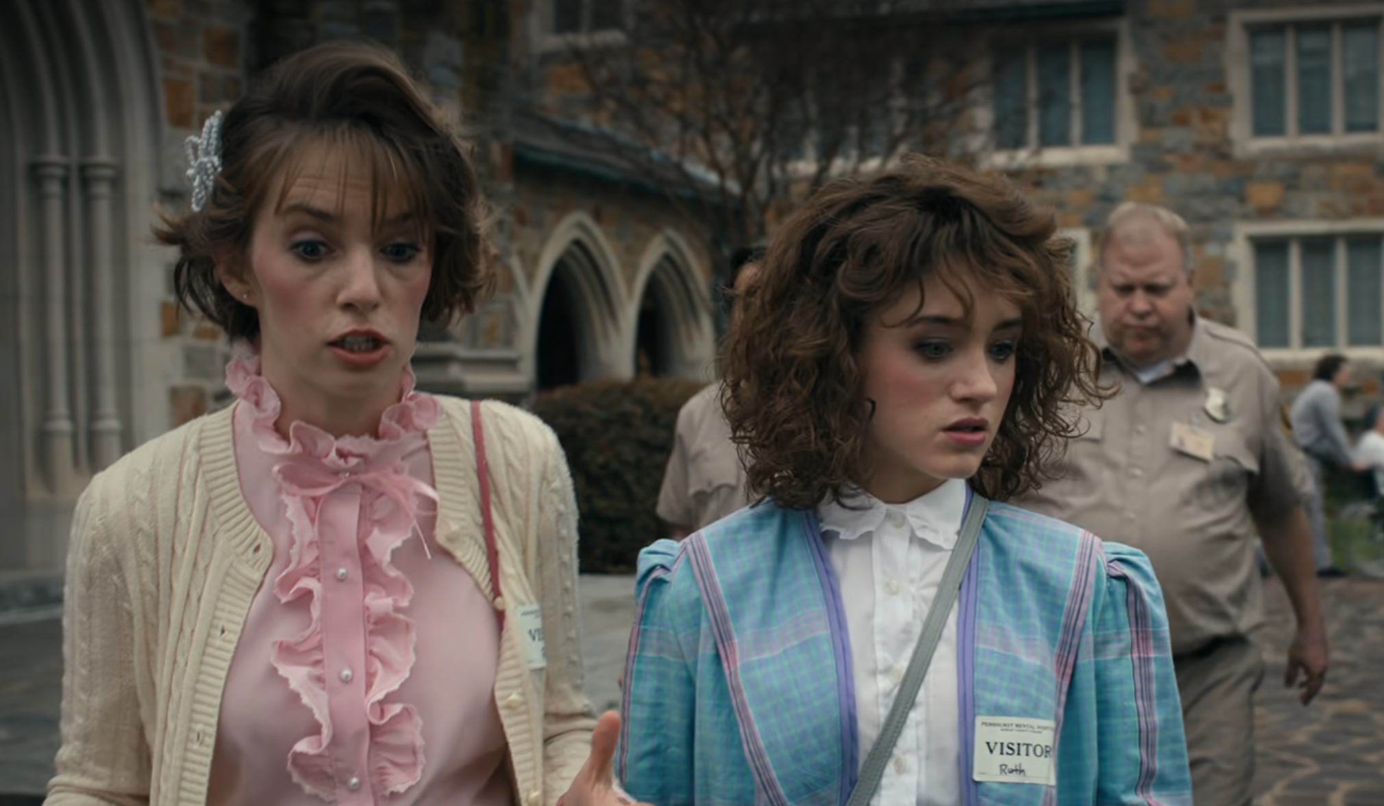 Two women in vintage-inspired outfits walk outside a stone building. They appear surprised or concerned. A man in uniform walks behind them