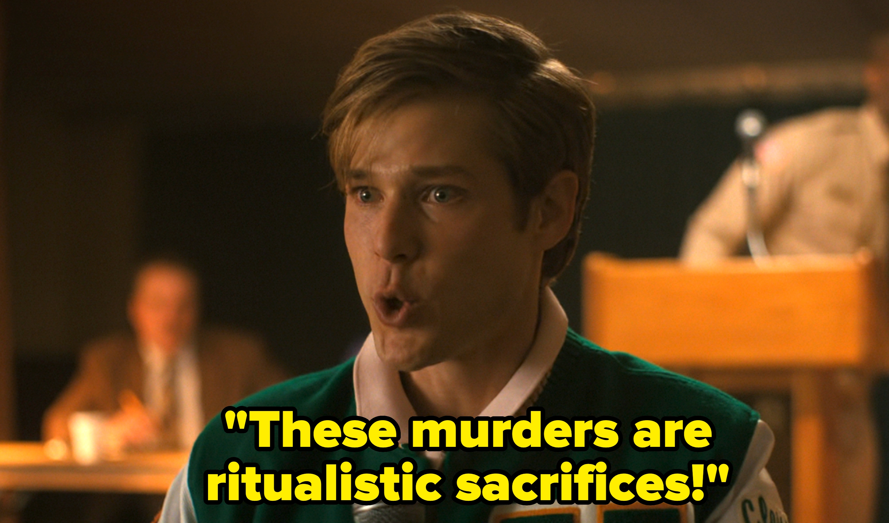 Person speaking with a surprised expression, holding a microphone, wearing a varsity-style jacket. Background shows blurred figure at a podium