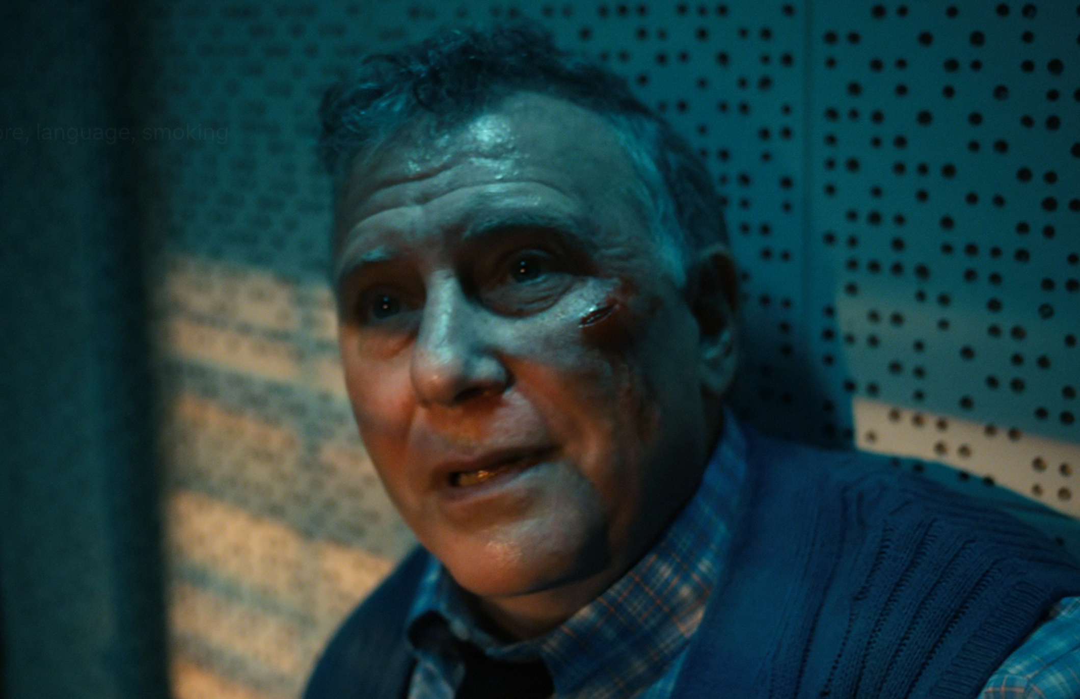 A man sits against a wall with a weary expression, showing a bruise on his cheek, under dramatic lighting