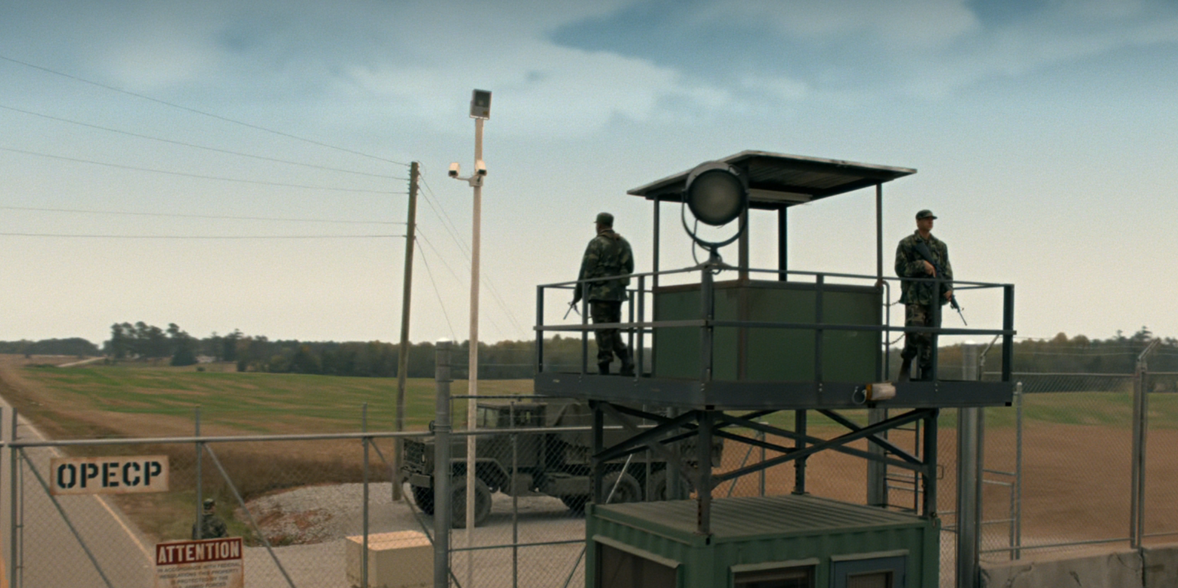 Two soldiers stand on a guard tower overlooking a fenced perimeter with fields in the background