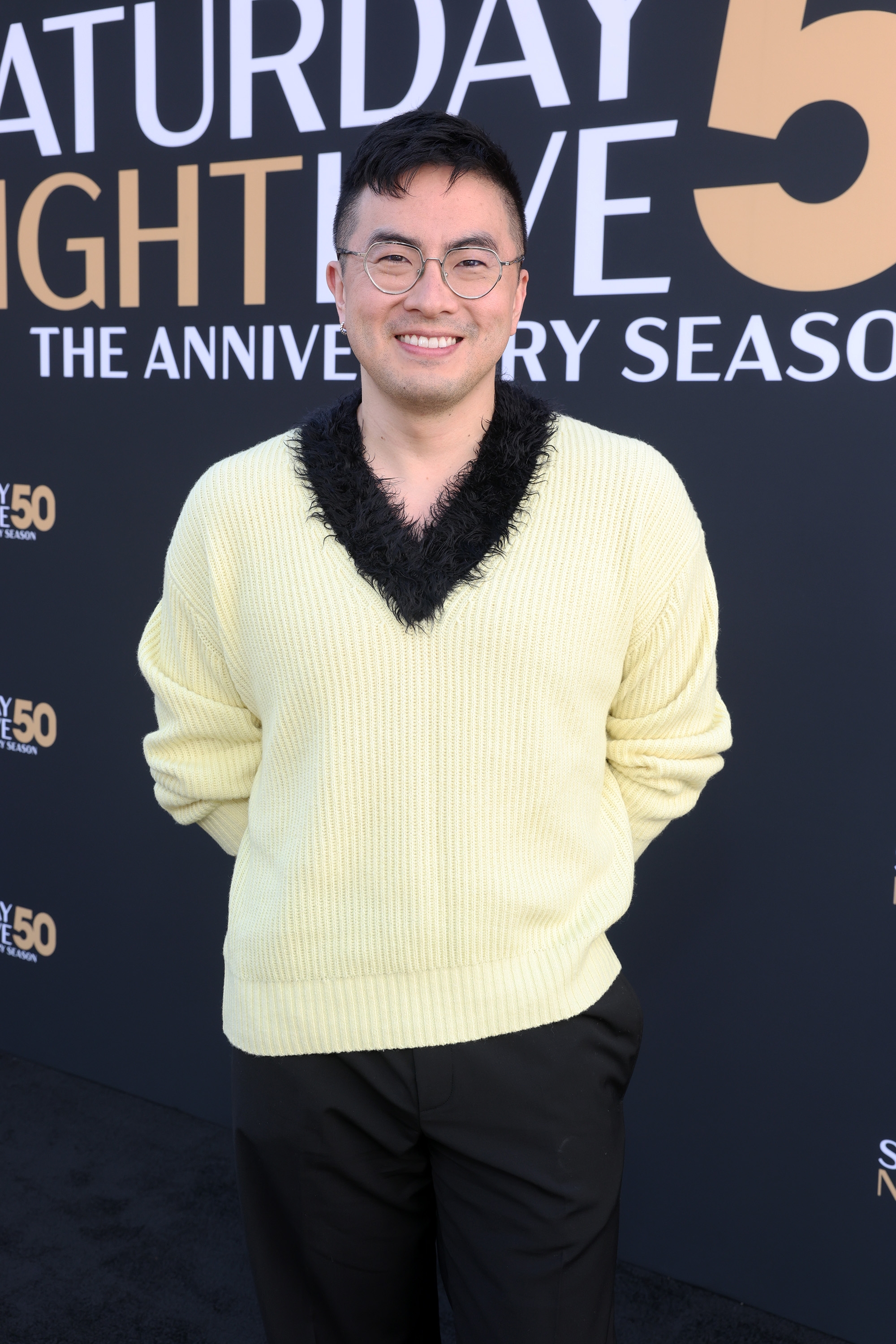 Person in a yellow sweater with black trim stands smiling at an event backdrop for a show's anniversary season