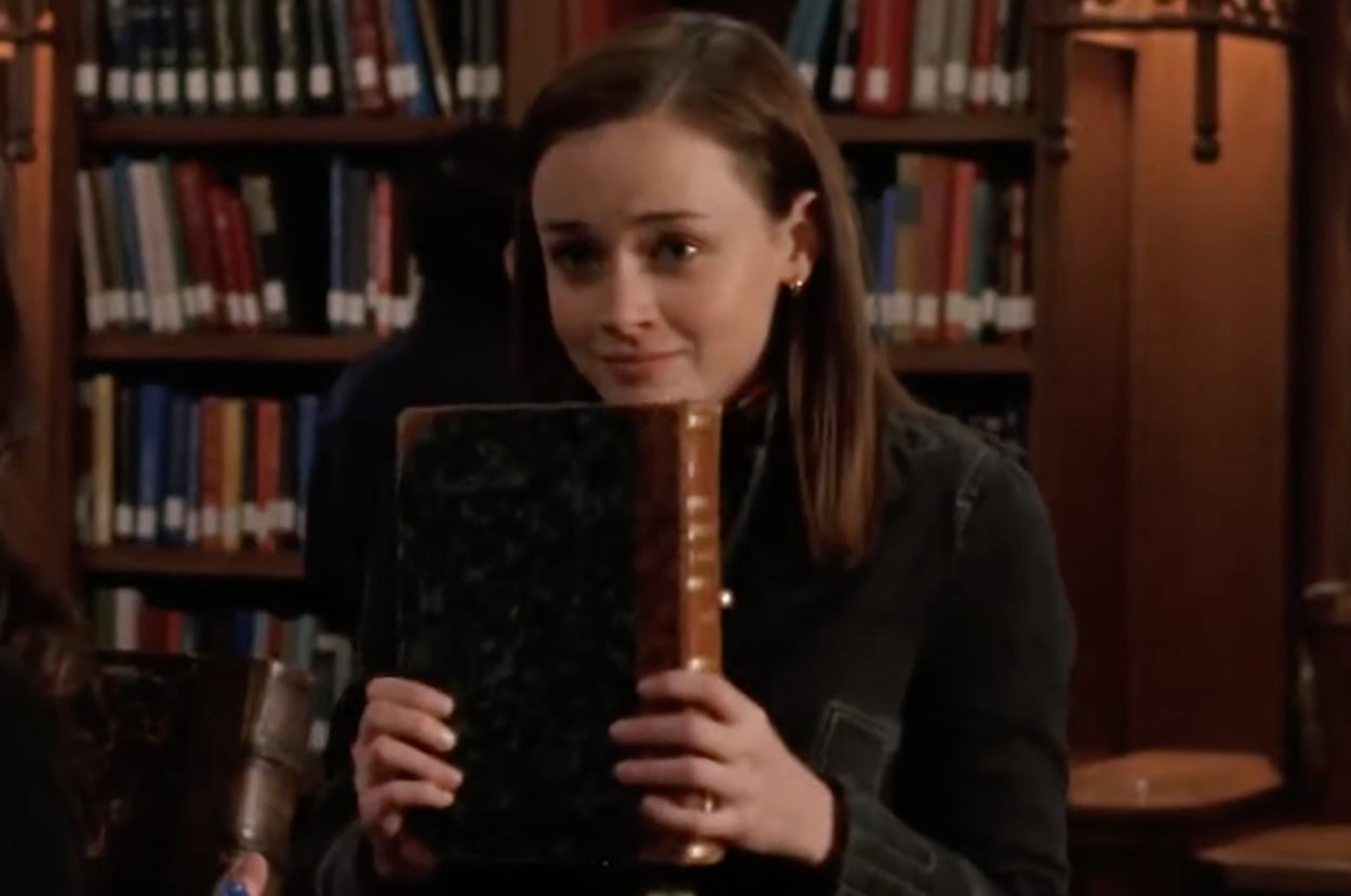 Rory Gilmore from "Gilmore Girls" holding a large book, smiling slightly with a thoughtful expression in a library setting. Bookshelves visible in the background