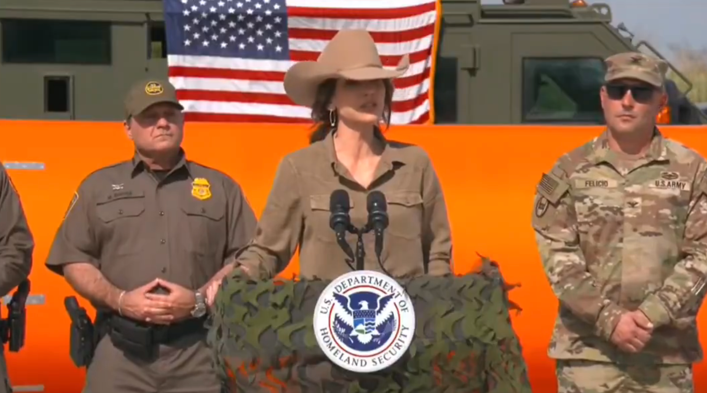 Offentlig person i en cowboyhatt som snakker på et podium med et emblem fra Dept. of Homeland Security, flankert av uniformert personell, amerikansk flagg i bakgrunnen