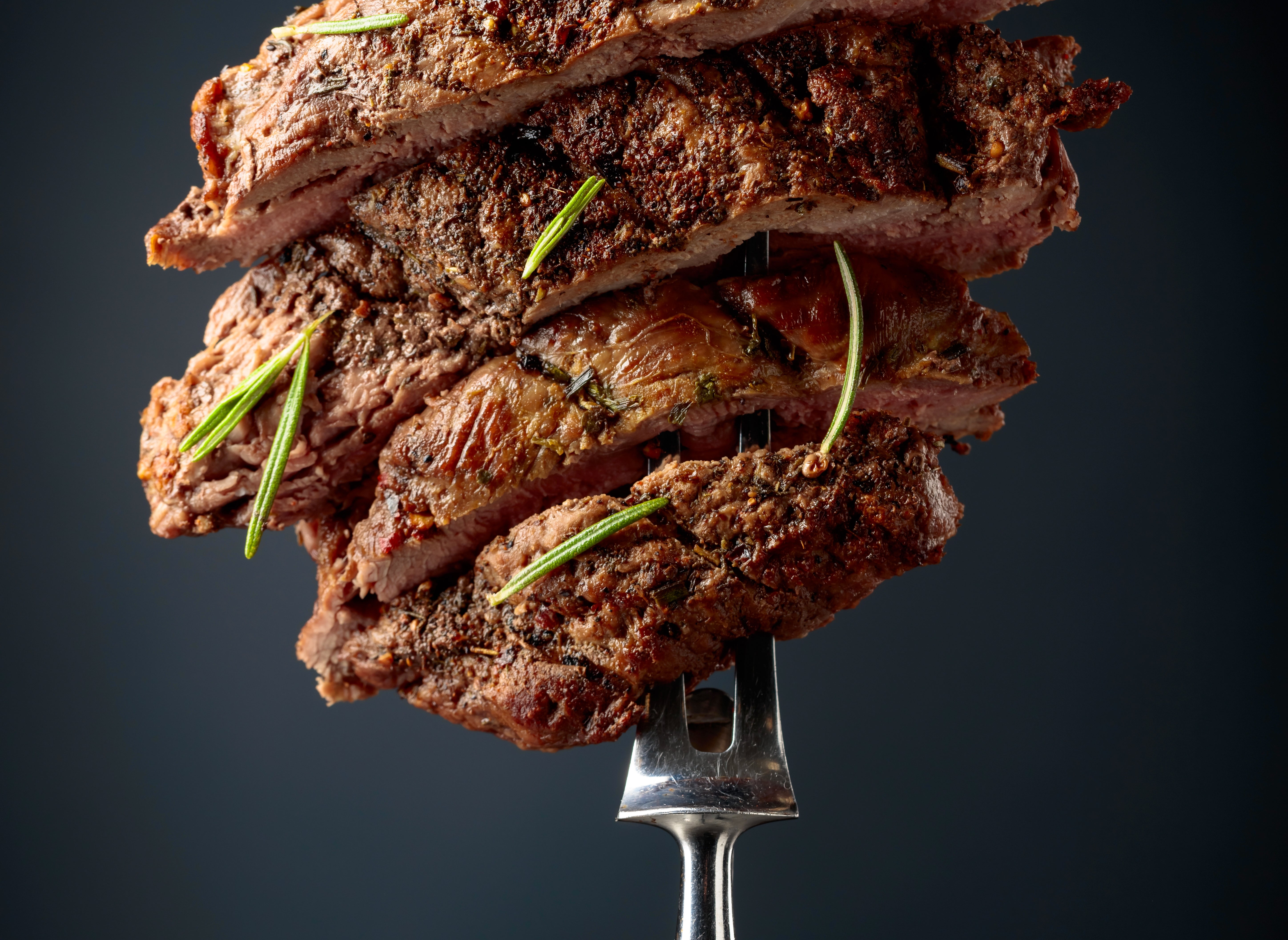 Slices of grilled steak stacked on a fork, garnished with rosemary sprigs, on a dark background