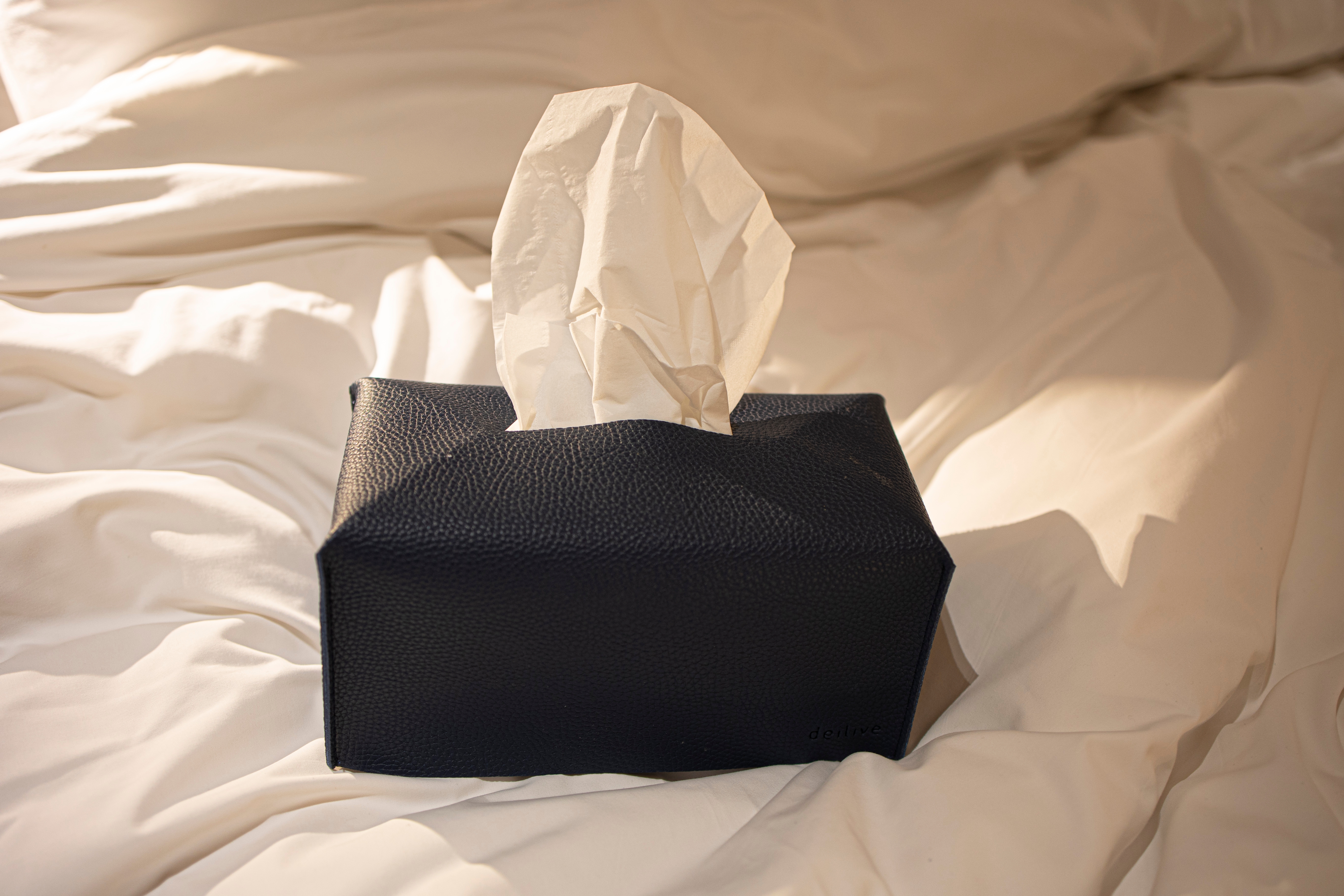 A leather tissue box with a tissue sticking out is placed on a rumpled bed, suggesting comfort or a home setting
