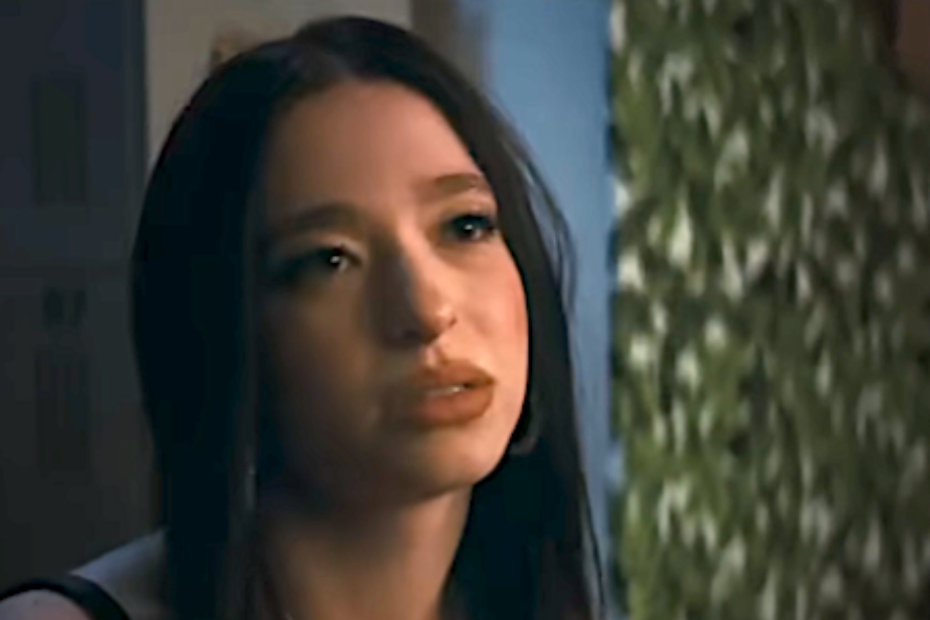 A person with long hair and a focused expression looks upward, set against a backdrop of greenery