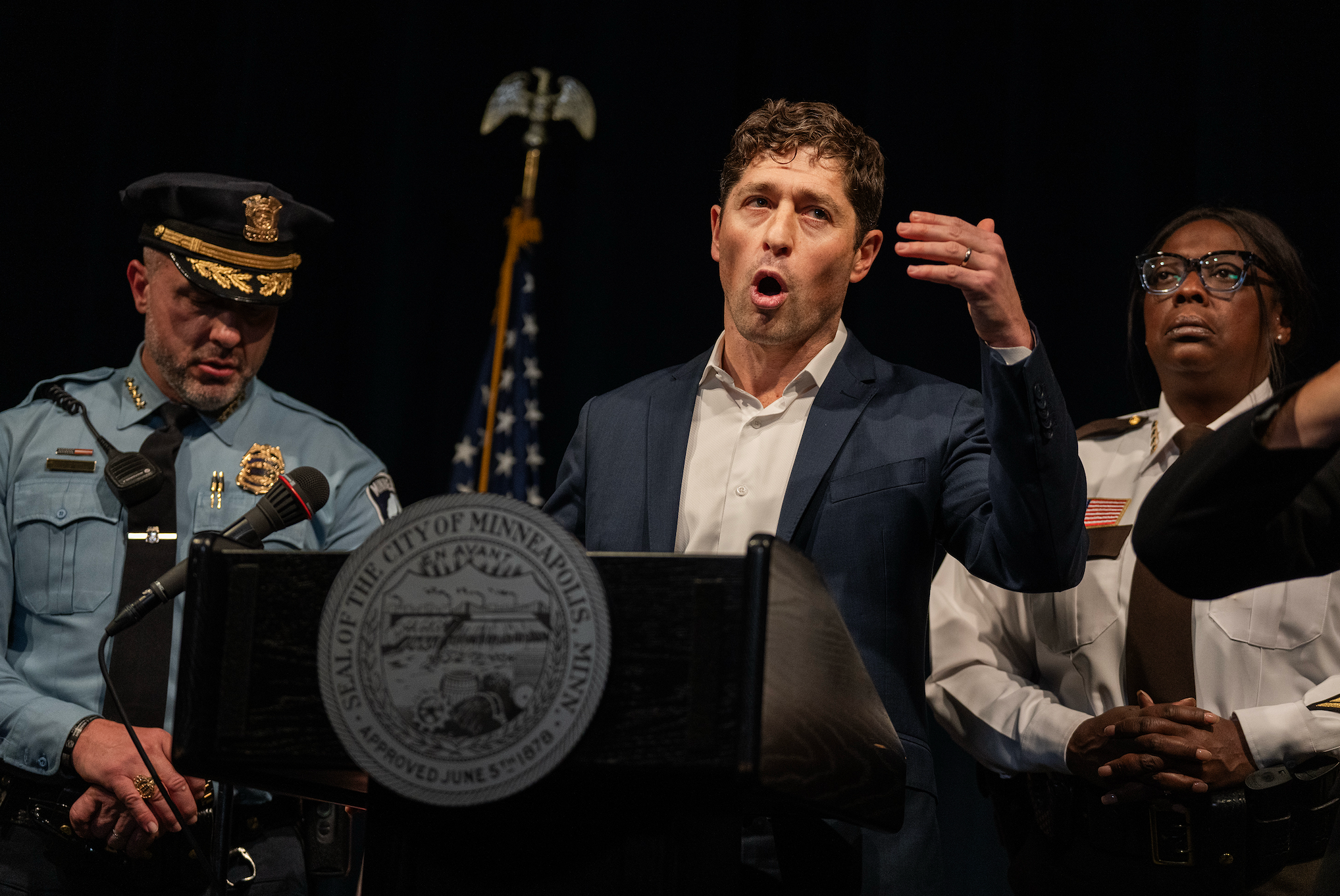 Ordfører Jacob Frey ved et podium gester under en pressekonferanse, flankert av to uniformerte offiserer, en i politiuniform og en i kjoleuniform