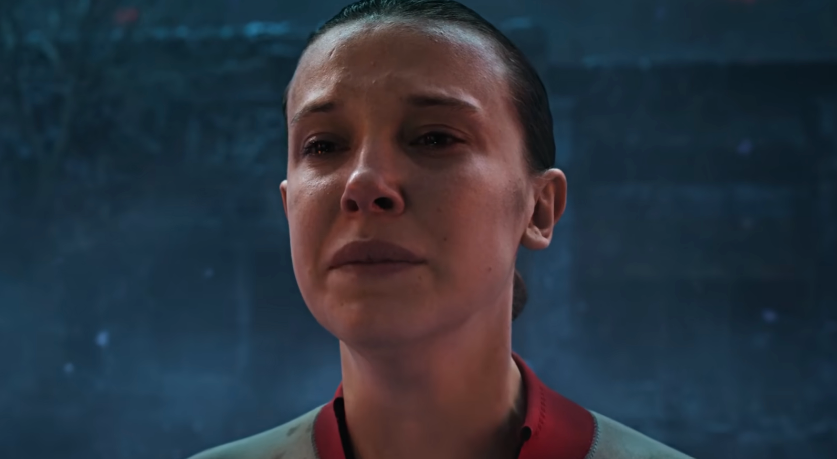 A person gazing upwards with a somber expression, short hair slicked back, wearing a light-colored shirt with a round collar in a dimly lit setting