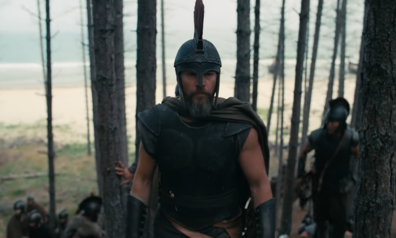 Man in ancient warrior attire with a helmet and armor walks through a forest, followed by others. Beach visible in the background