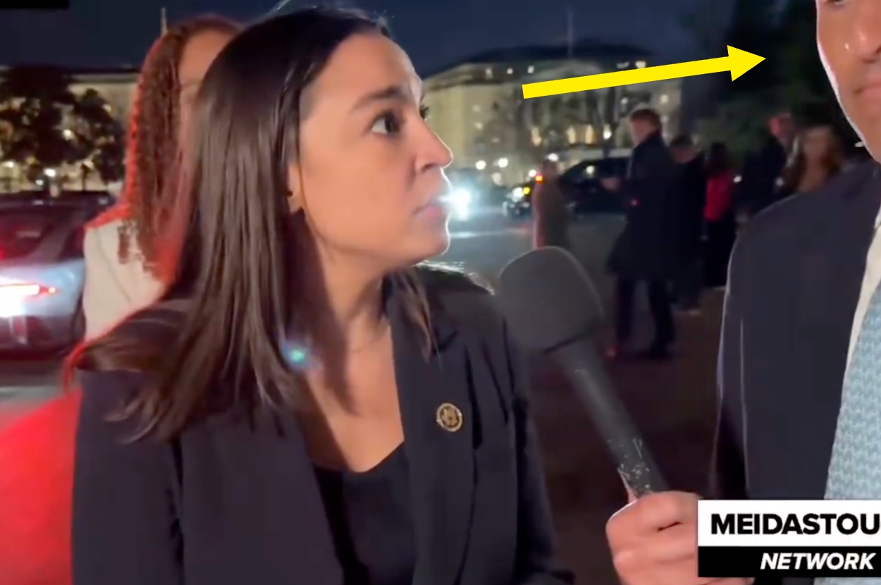 A person wearing formal attire speaks into a microphone during an outdoor event at night, with a building visible in the background