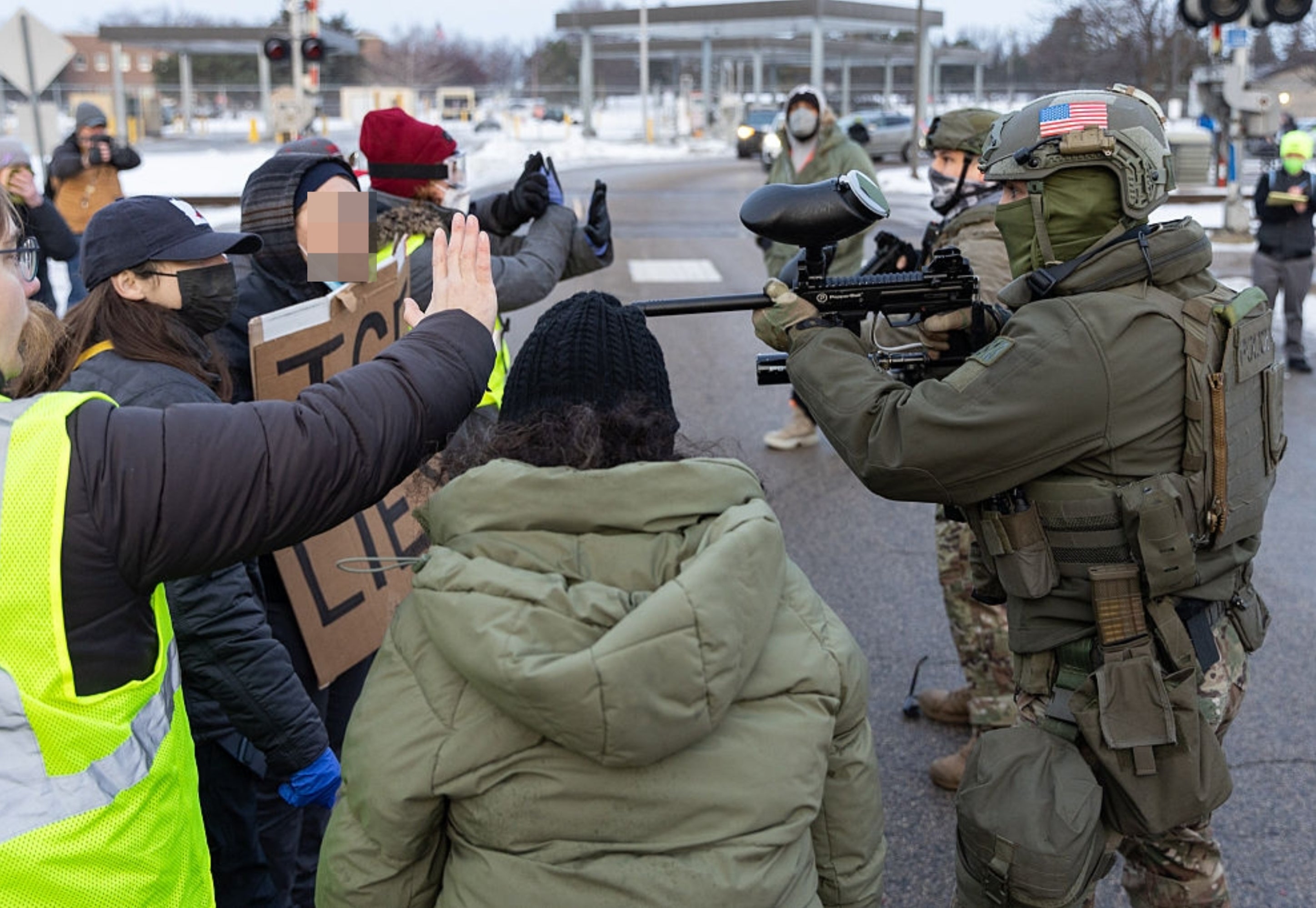 En soldat i taktisk utstyr konfronterer en gruppe demonstranter under en demonstrasjon, en holder et skilt, i en bygate. Spenningen er synlig