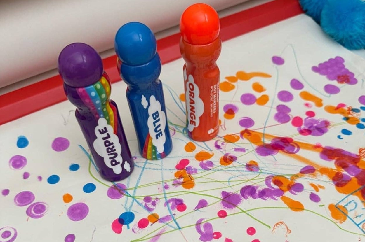 Three bingo daubers labeled purple, blue, and orange stand upright on a table with colorful spots and lines scattered on a sheet of paper