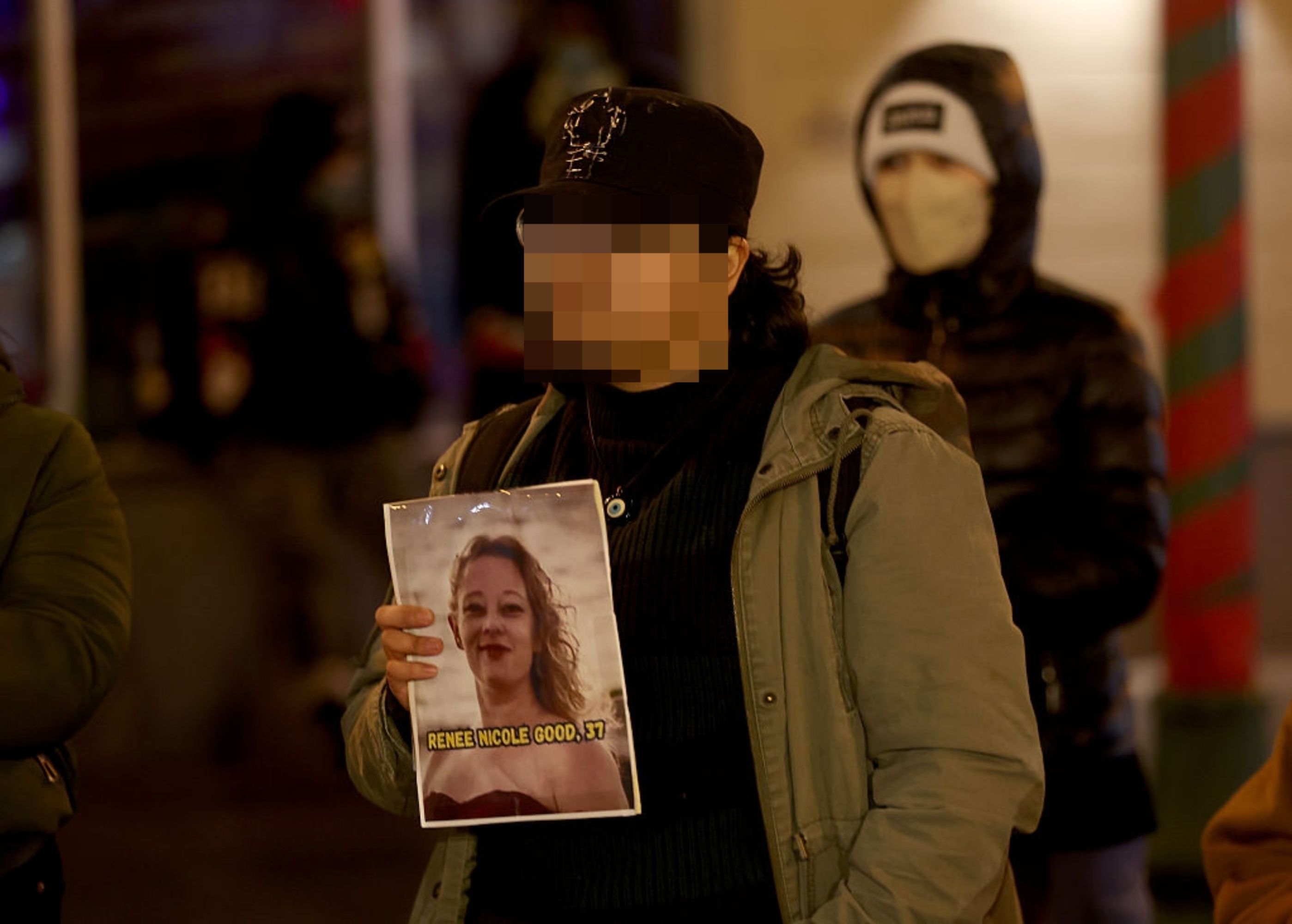 Person som holder et bilde av en kvinne med "RENEE NICOLE GOOD 37" trykt på den, stående utendørs blant andre