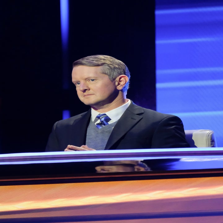 Person on a TV game show set wearing a suit and tie, seated behind a desk, focused and attentive