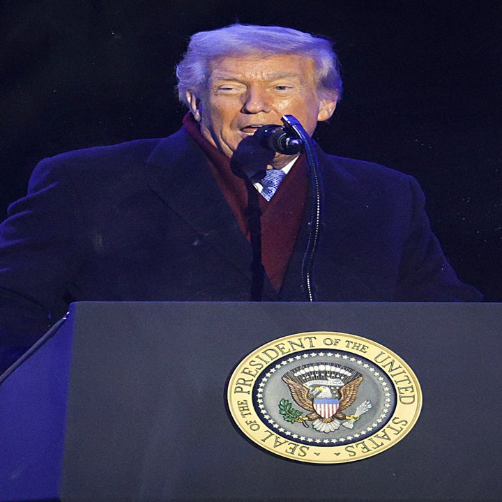 A man standing at a podium with the U.S. Presidential seal, speaking into a microphone while wearing a dark coat at an event