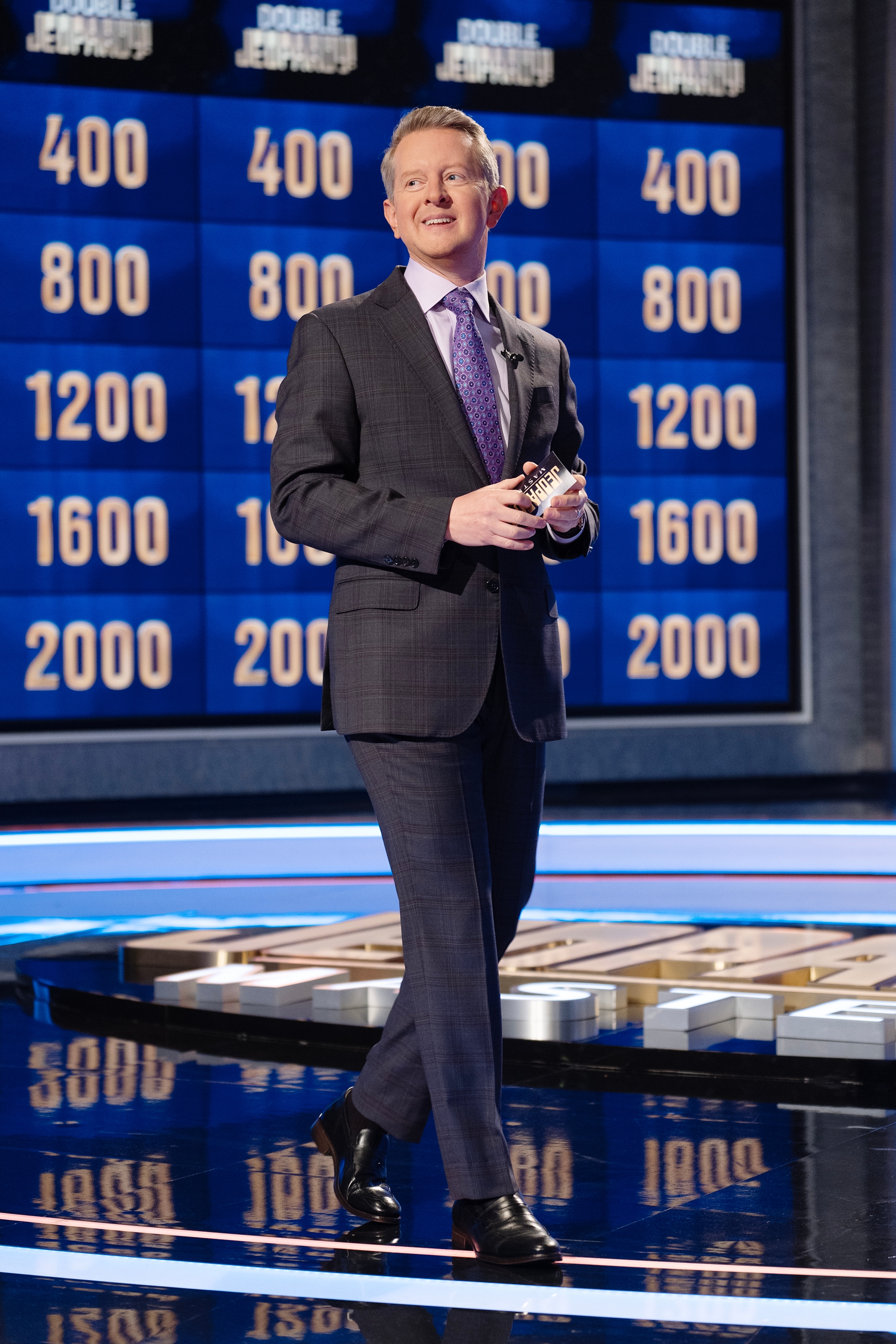 Ken Jennings in a checkered suit holds a card on a game show set with a board featuring numbers in the background
