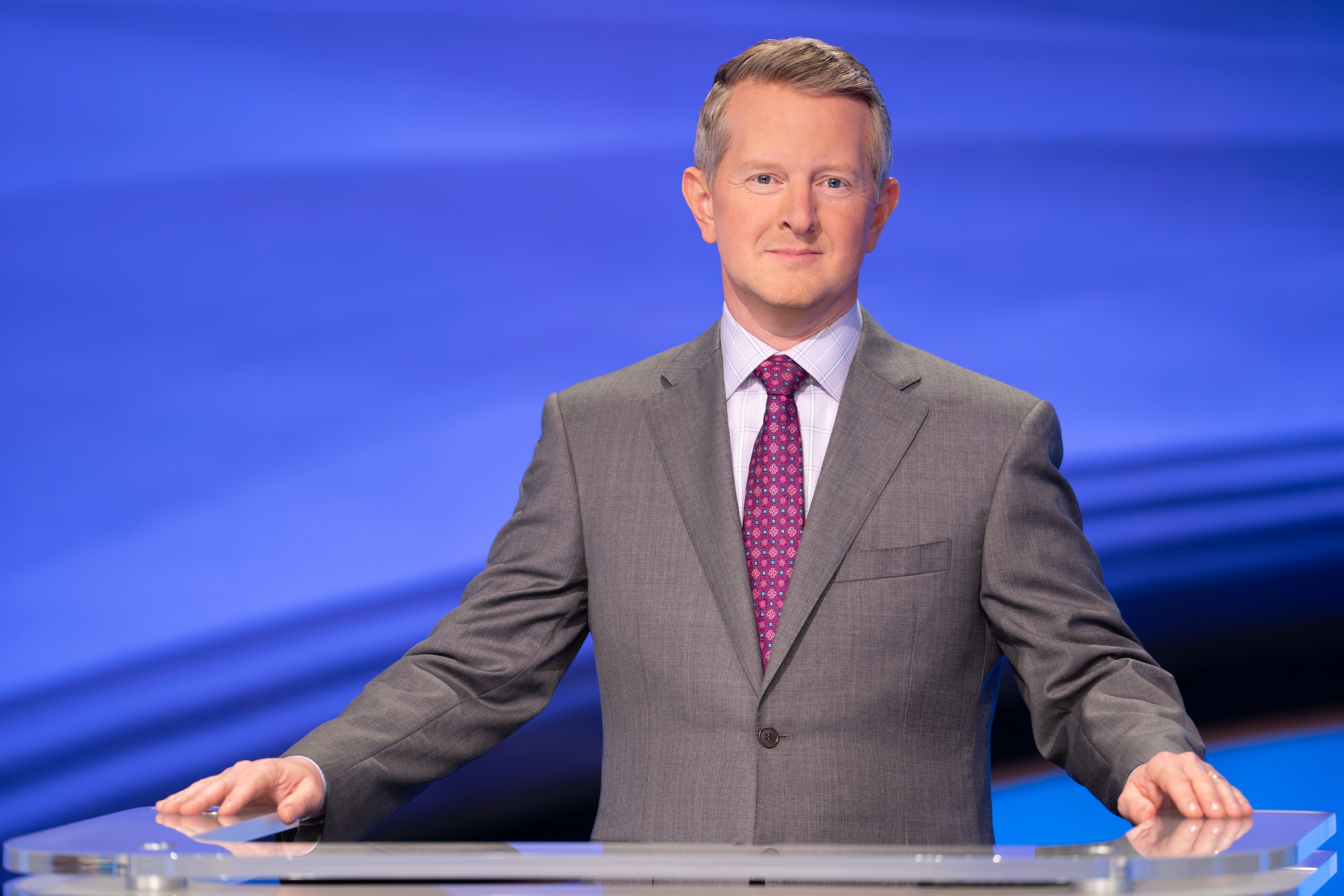 Ken Jennings in a suit stands at the Jeopardy podium, facing the camera with a confident expression