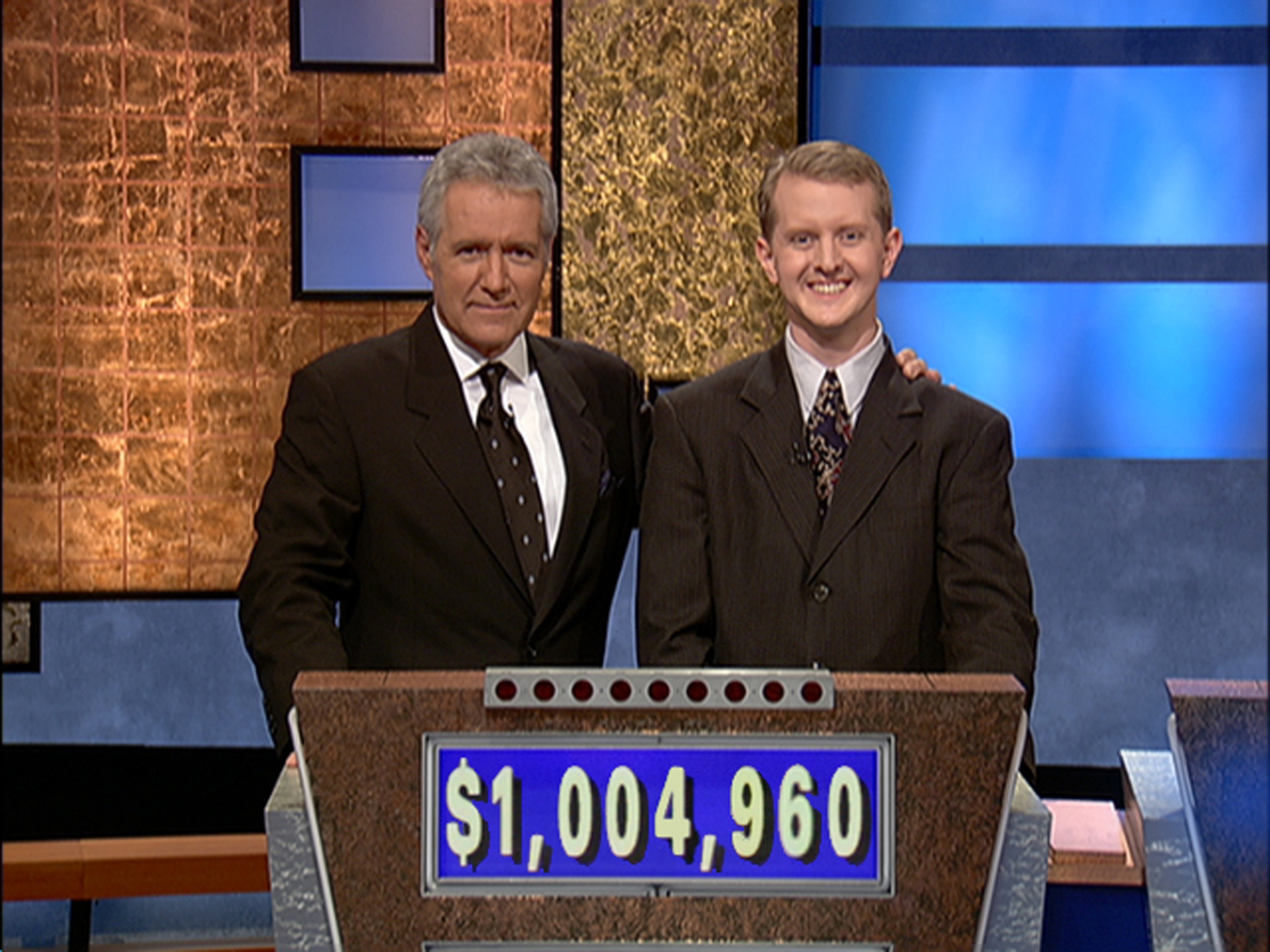 Alex Trebek and Ken Jennings in suits stand on the Jeopardy set with Alex holding Ken's shoulder