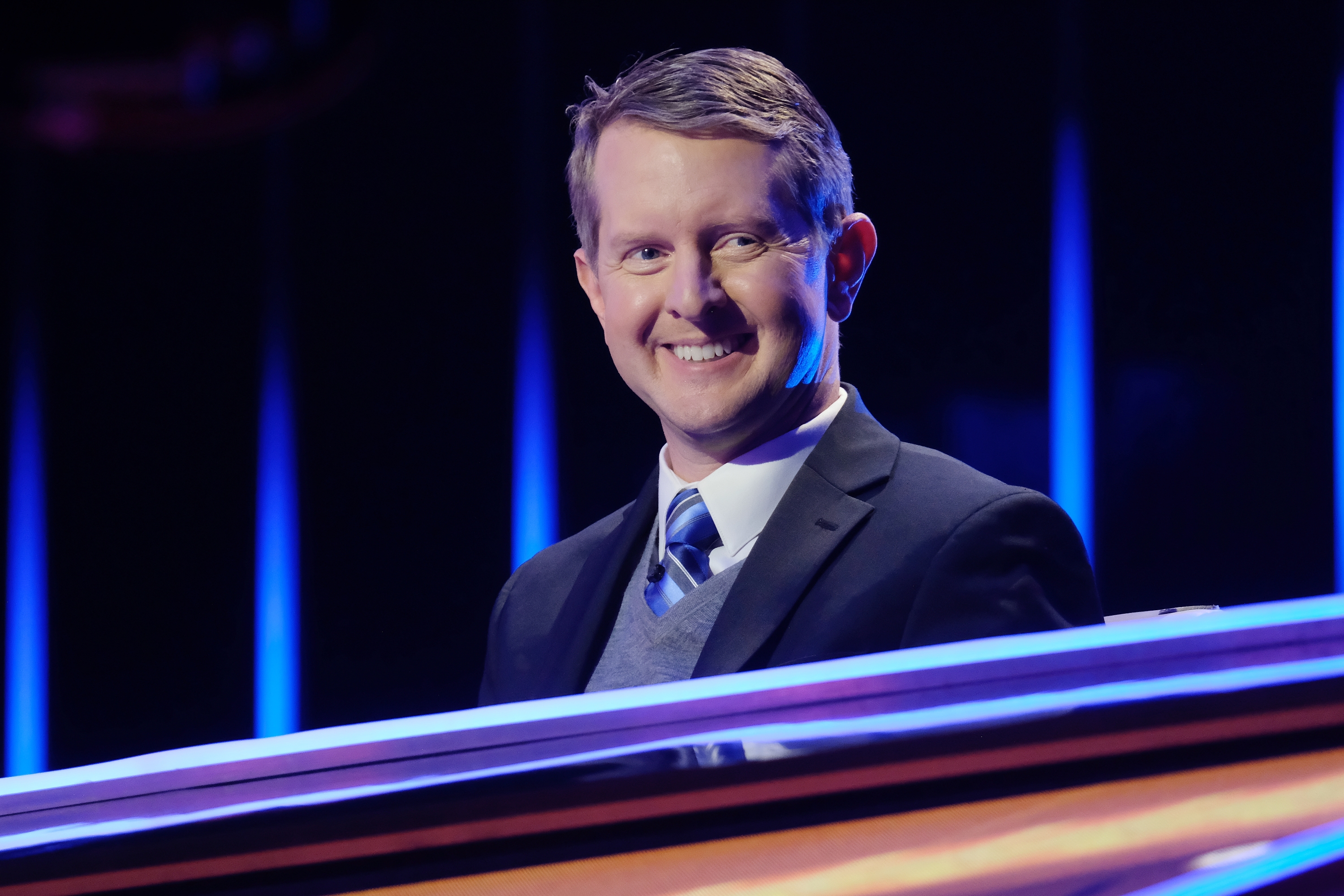 Ken Jennings in a suit and tie stands behind a game show podium smiling
