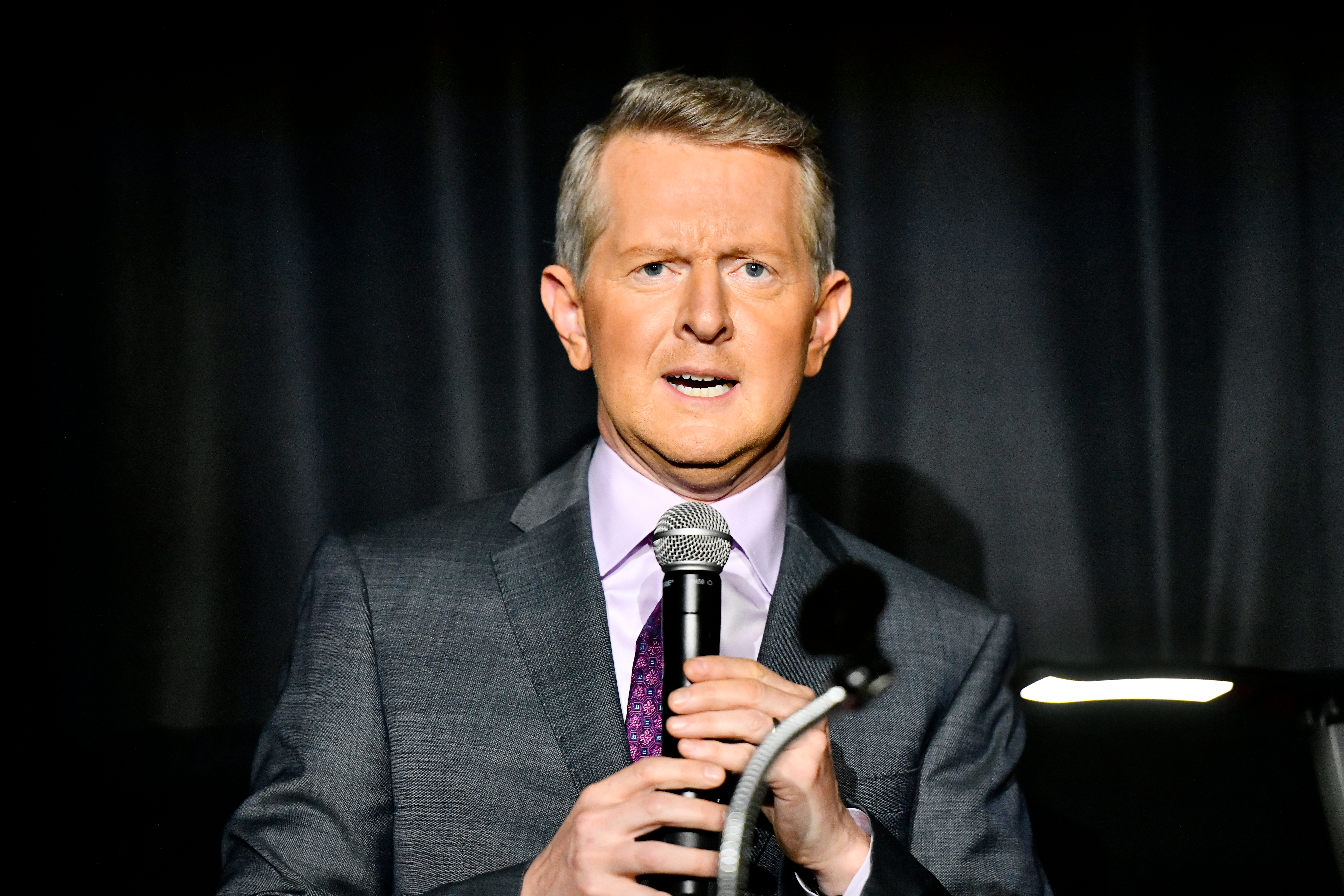 Ken Jennings in a suit holding a microphone, appearing to be speaking or presenting on a stage with a dark background