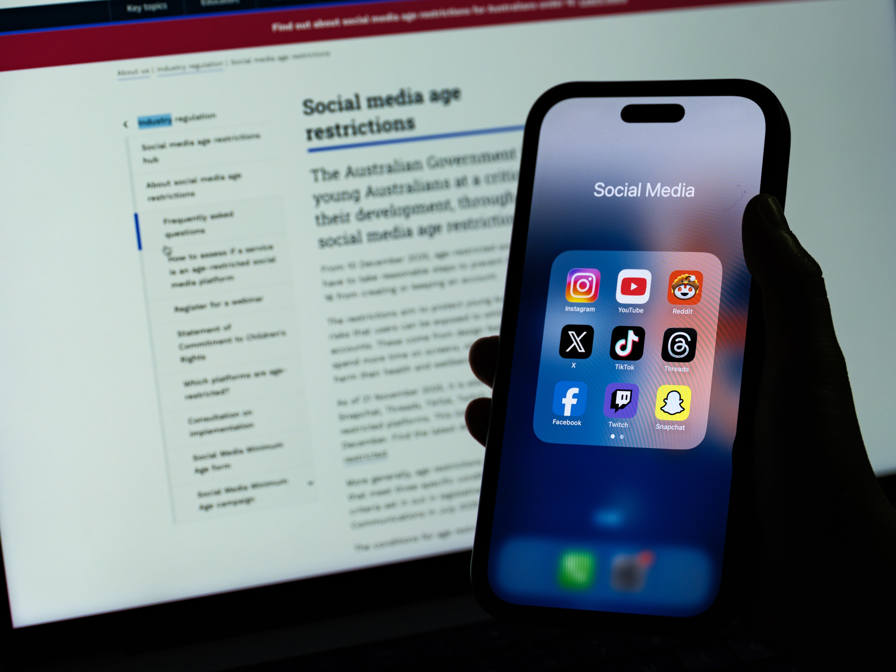 Telefonskjerm som viser apper for sosiale medier holdt foran en datamaskin som viser tekst om aldersbegrensninger på sosiale medier