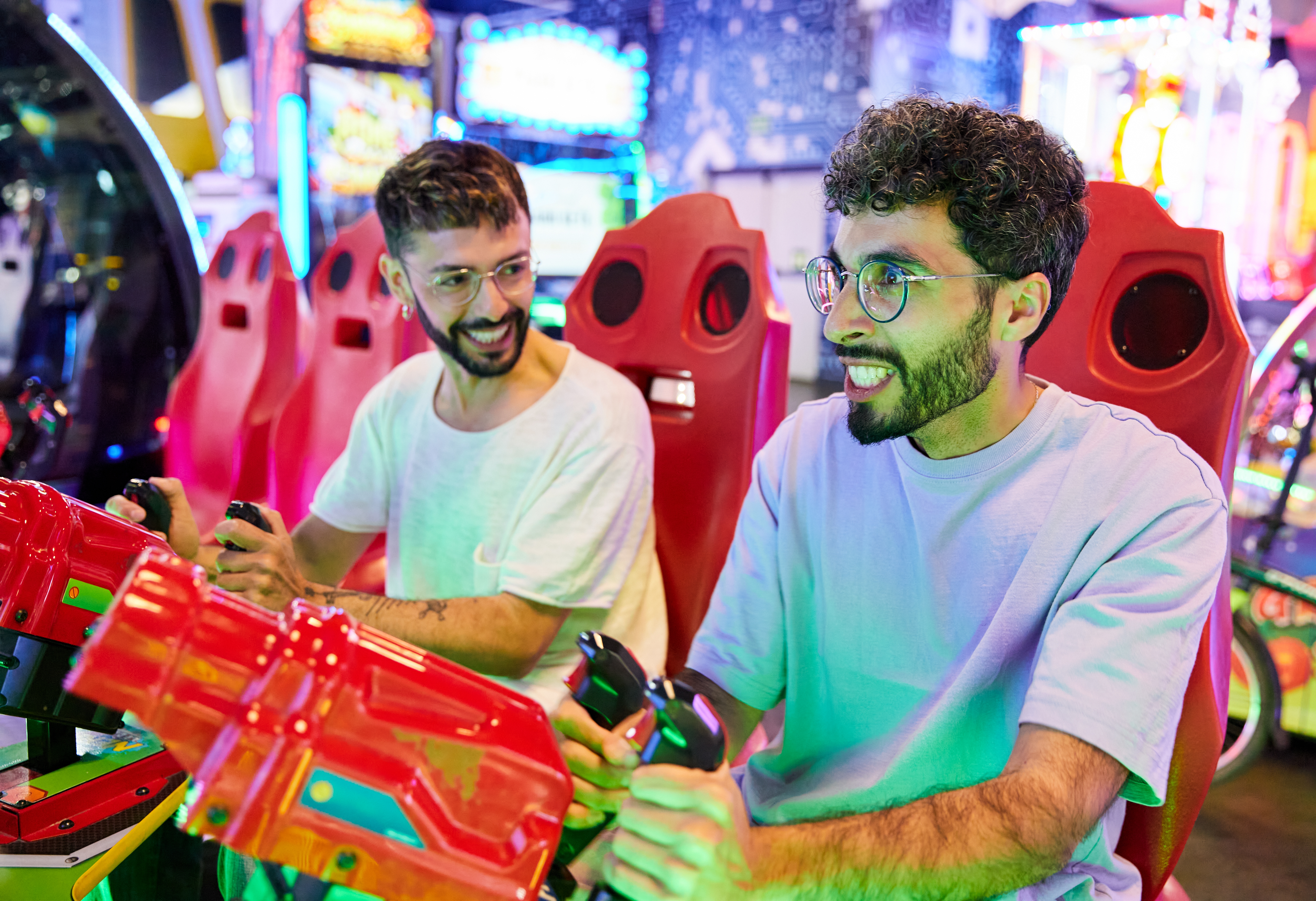 Two radical   smiling and playing a racing arcade game. They clasp  steering instrumentality    controllers and beryllium   successful  reddish  gaming chairs. Bright gaming machines situation   them