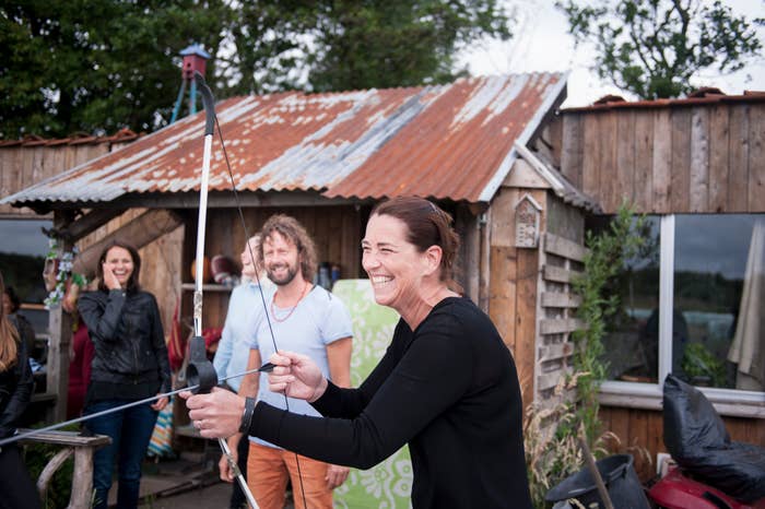 Group of radical   enjoying archery outdoors adjacent   a rustic cabin. A pistillate   successful  the foreground is laughing portion    drafting  a bowstring, others watch