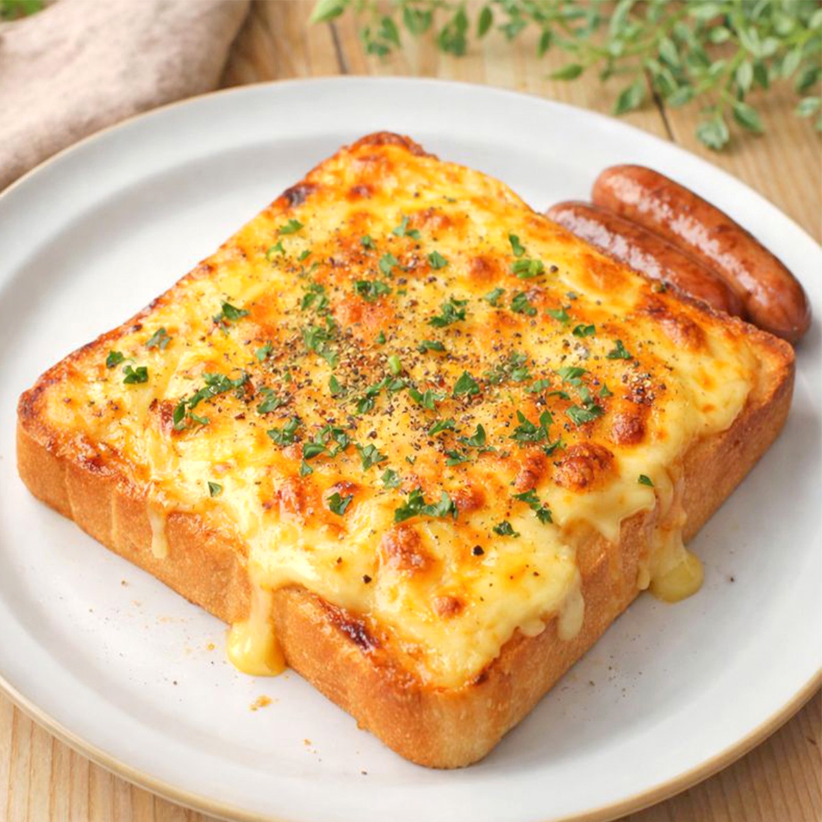 とろけるチーズとパセリがのったトーストが皿に盛られ、横にソーセージが添えられています。