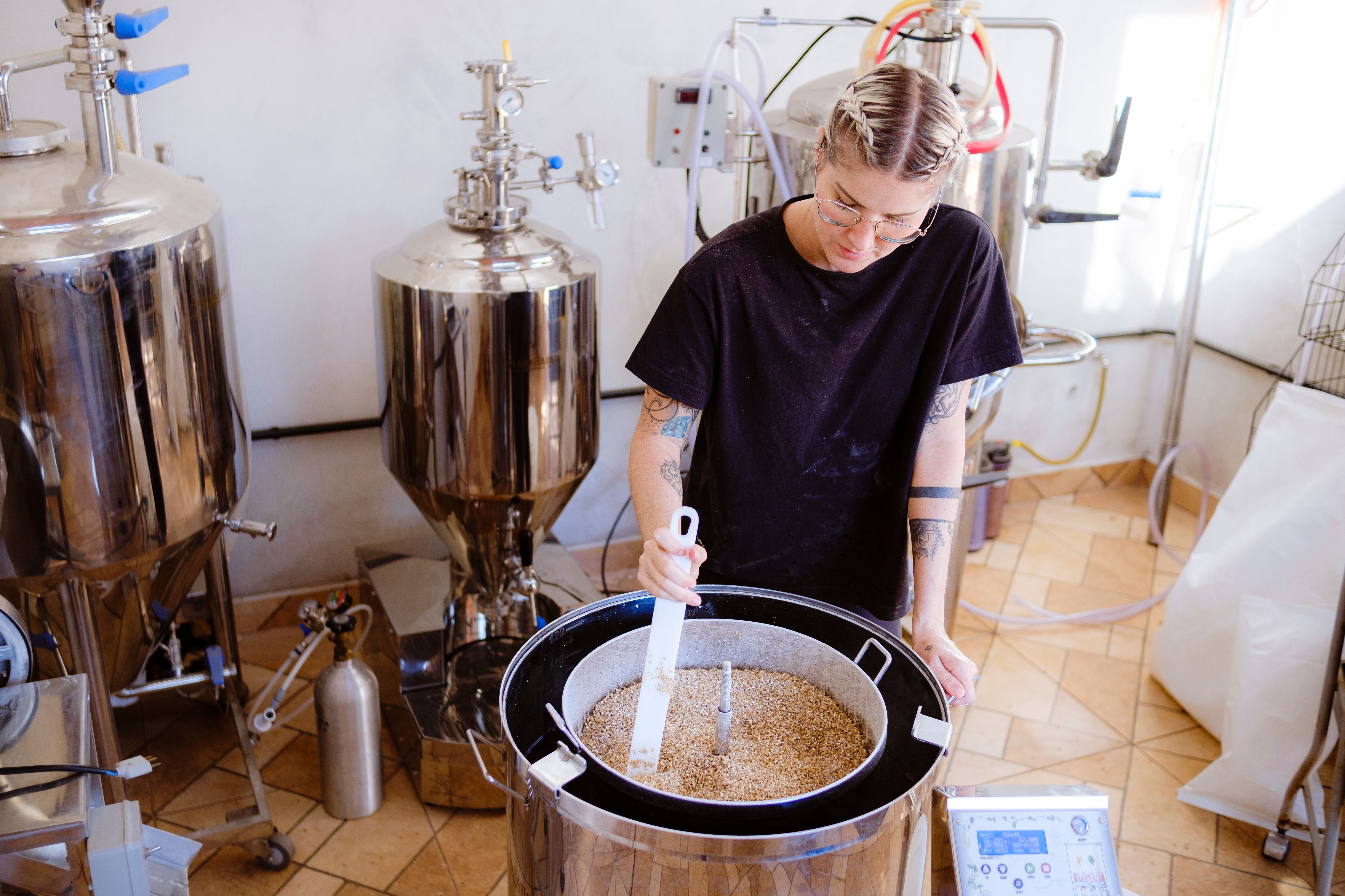 Person stirring grains successful  a brewery with fermentation tanks successful  the background. They're wearing a casual achromatic  garment  and person  disposable   limb  tattoos