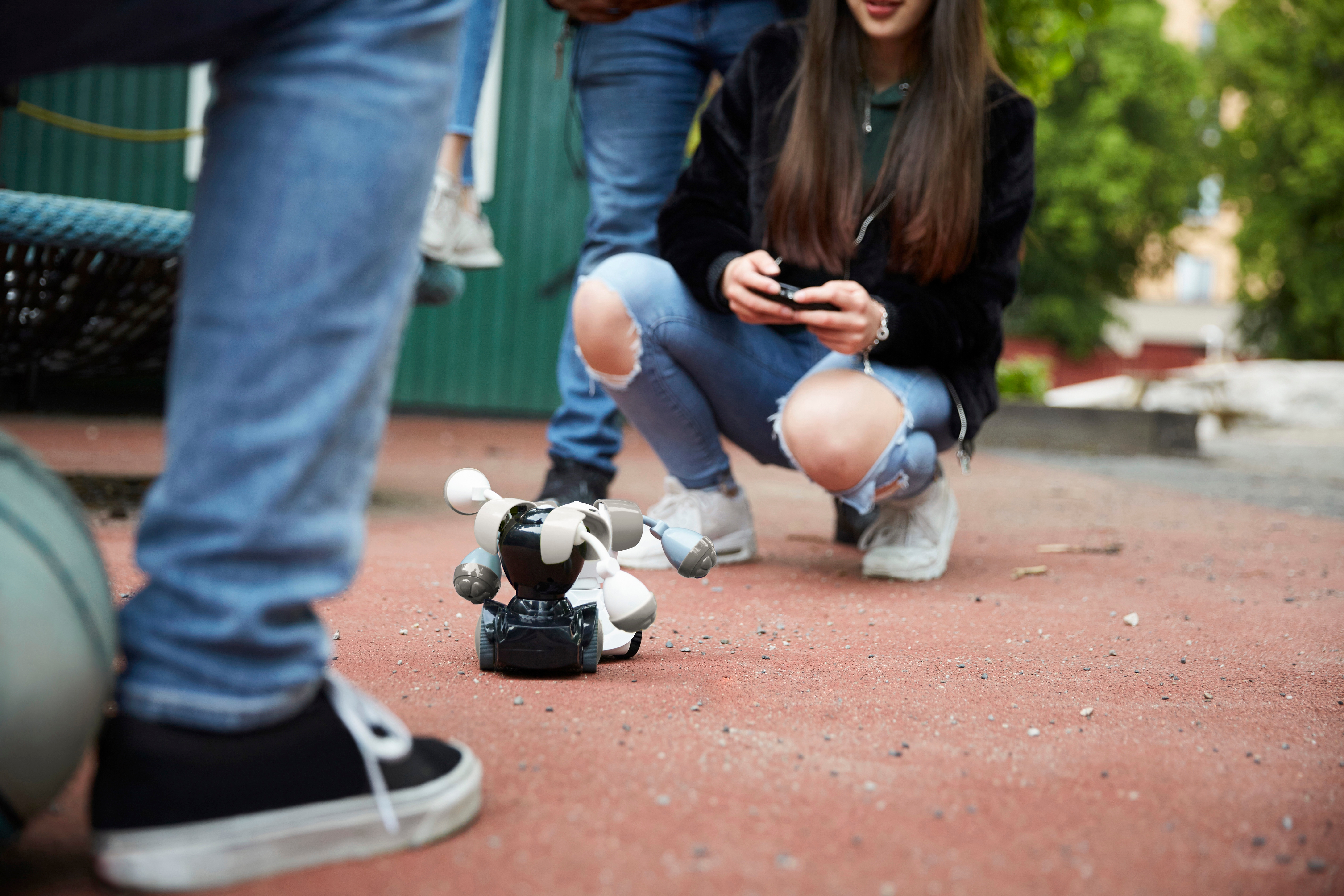 Person kneeling outdoors, utilizing a distant    power  to run  a tiny  robotic artifact  connected  the ground. Casual attire with jeans and sneakers