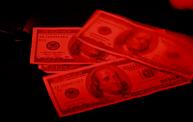 Several US hundred-dollar bills are spread out, partially obscured by red lighting