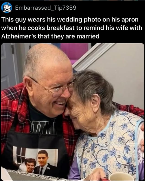 Elderly antheral   with an apron showing his wedding photo, hugging his wife, who has Alzheimer's. They are smiling astatine  a meal  table