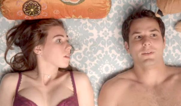A woman in a bra and a shirtless man lie on a bed, looking thoughtful. A round pillow with an ornate design is seen near their heads