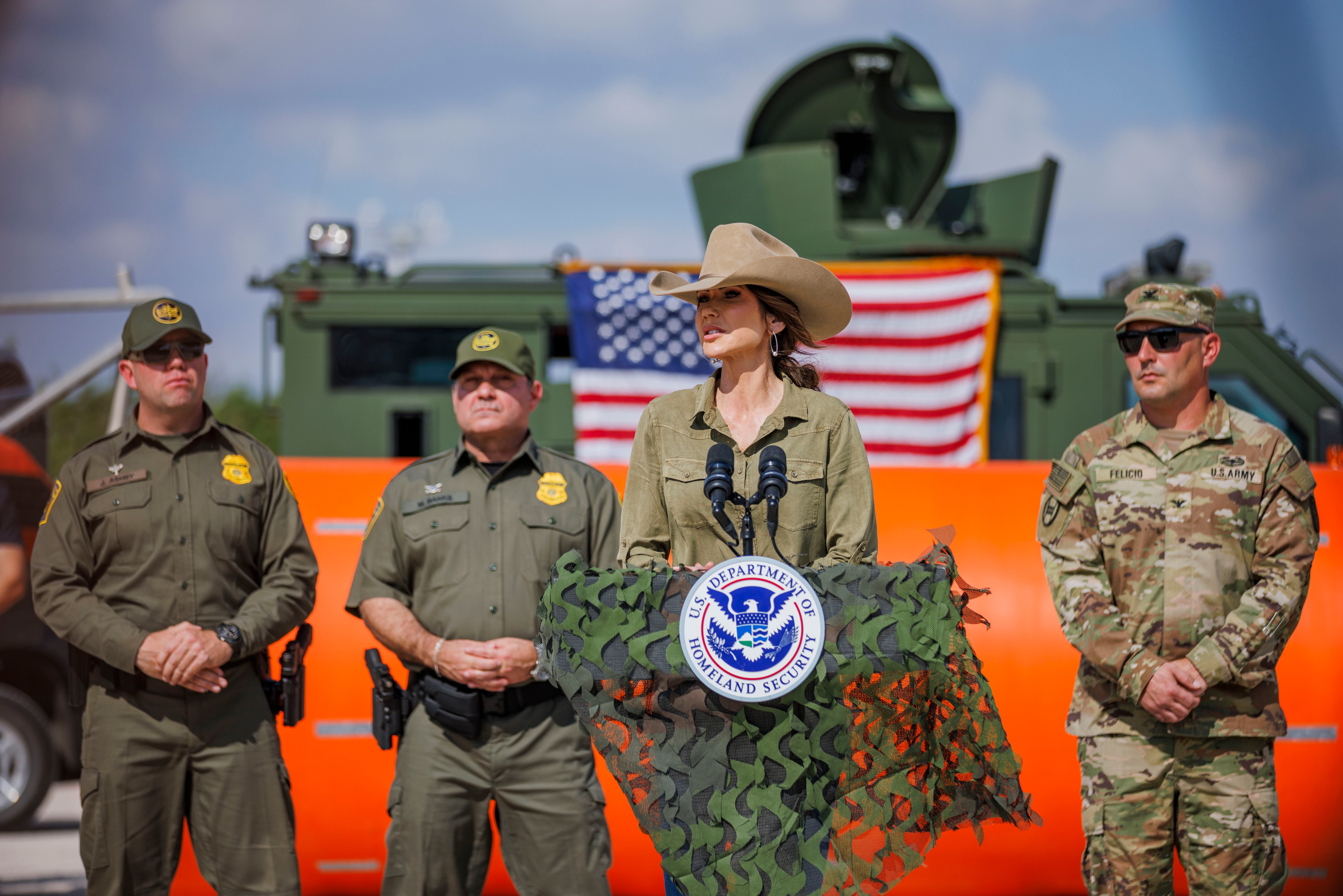 En person i cowboyhatt snakker ved et podium med et Homeland Security-emblem, flankert av uniformert personell og et amerikansk flagg i bakgrunnen