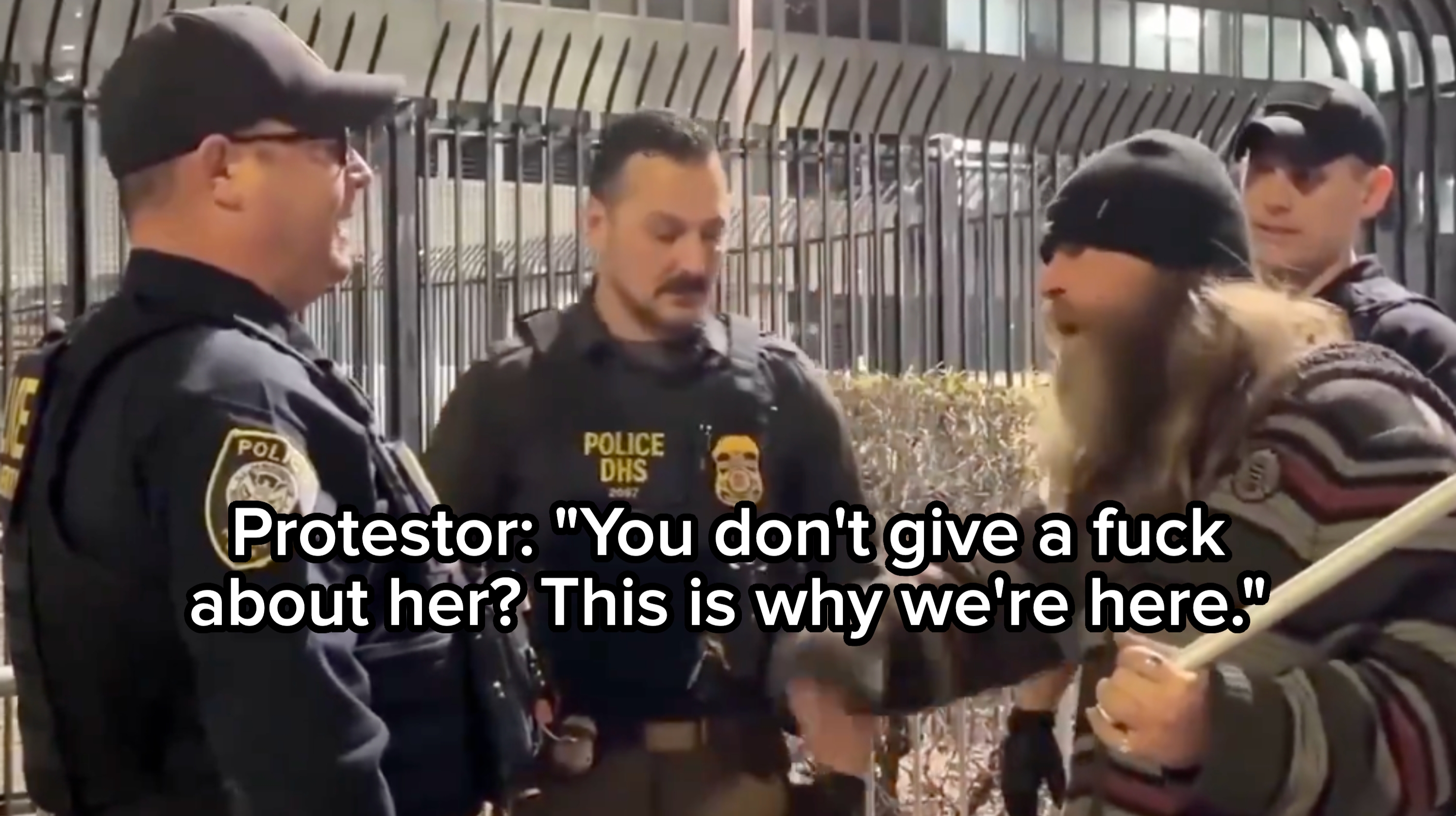 Tre politimenn går i samtale med en langhåret person som holder en pinne, utenfor en inngjerdet bygning