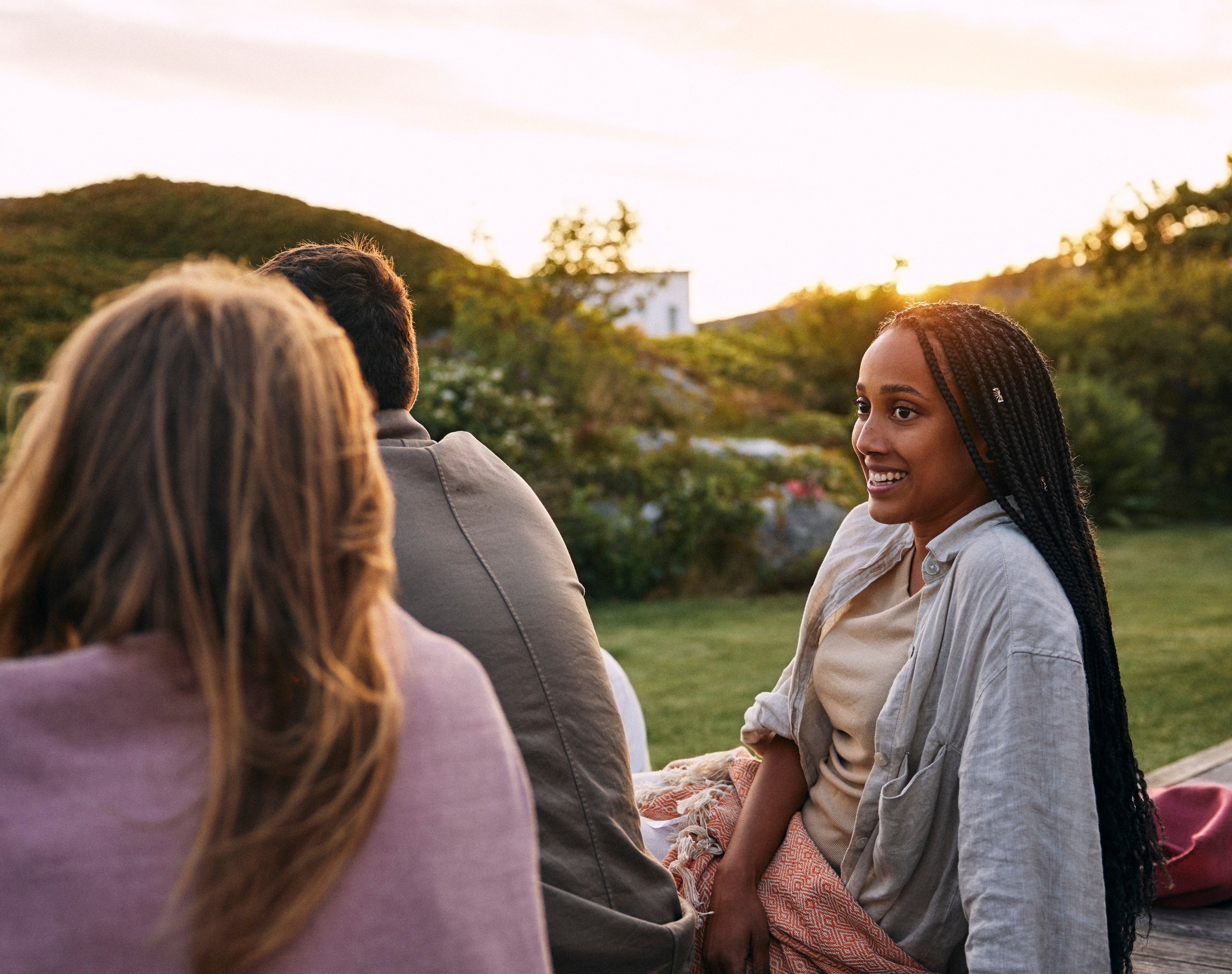 People sitting outdoors on a deck, engaged in conversation, with one person smiling and looking to the side, during a serene sunset
