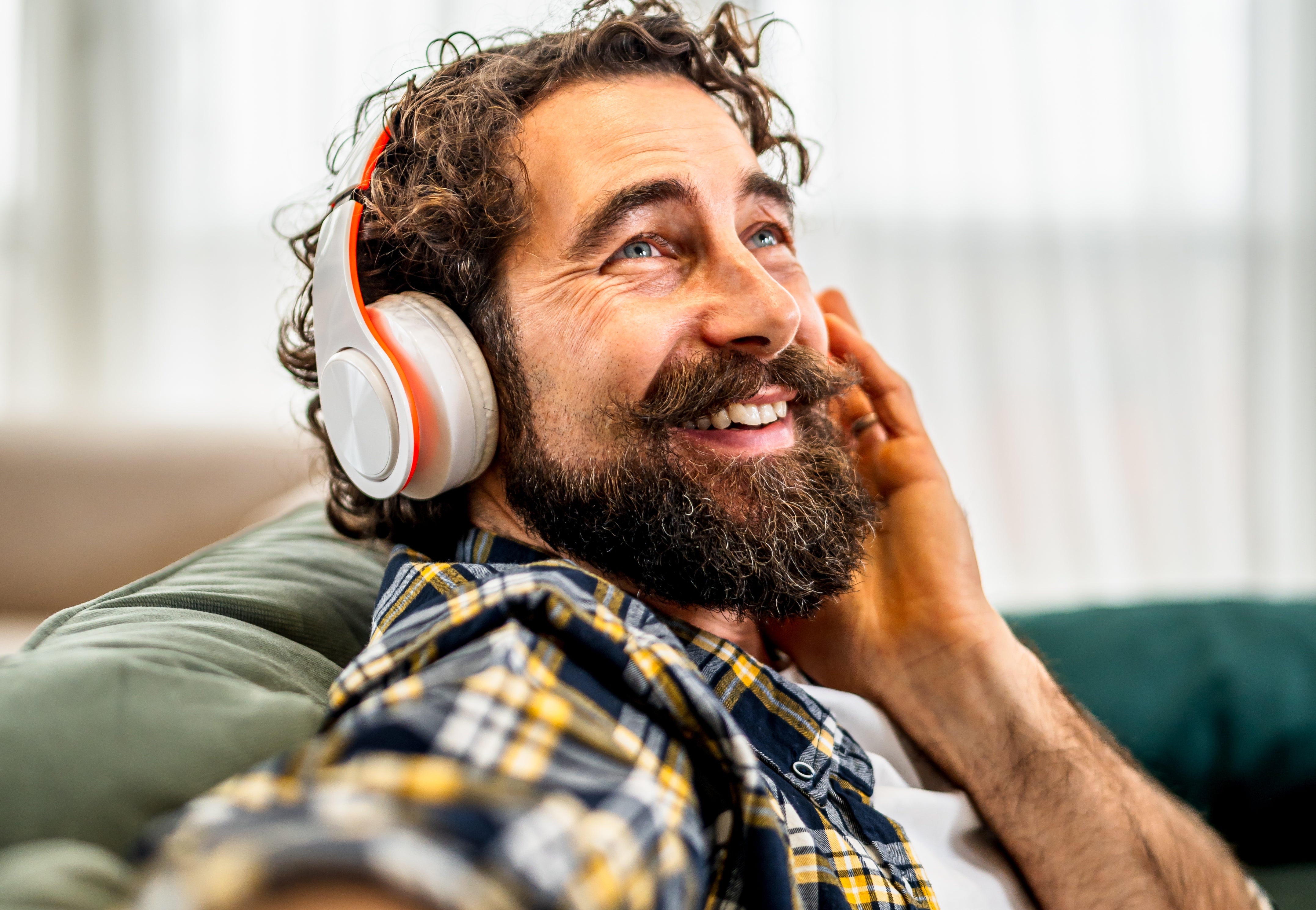 A bearded man wearing headphones smiles while taking a selfie, lounging casually in a plaid shirt over a white T-shirt