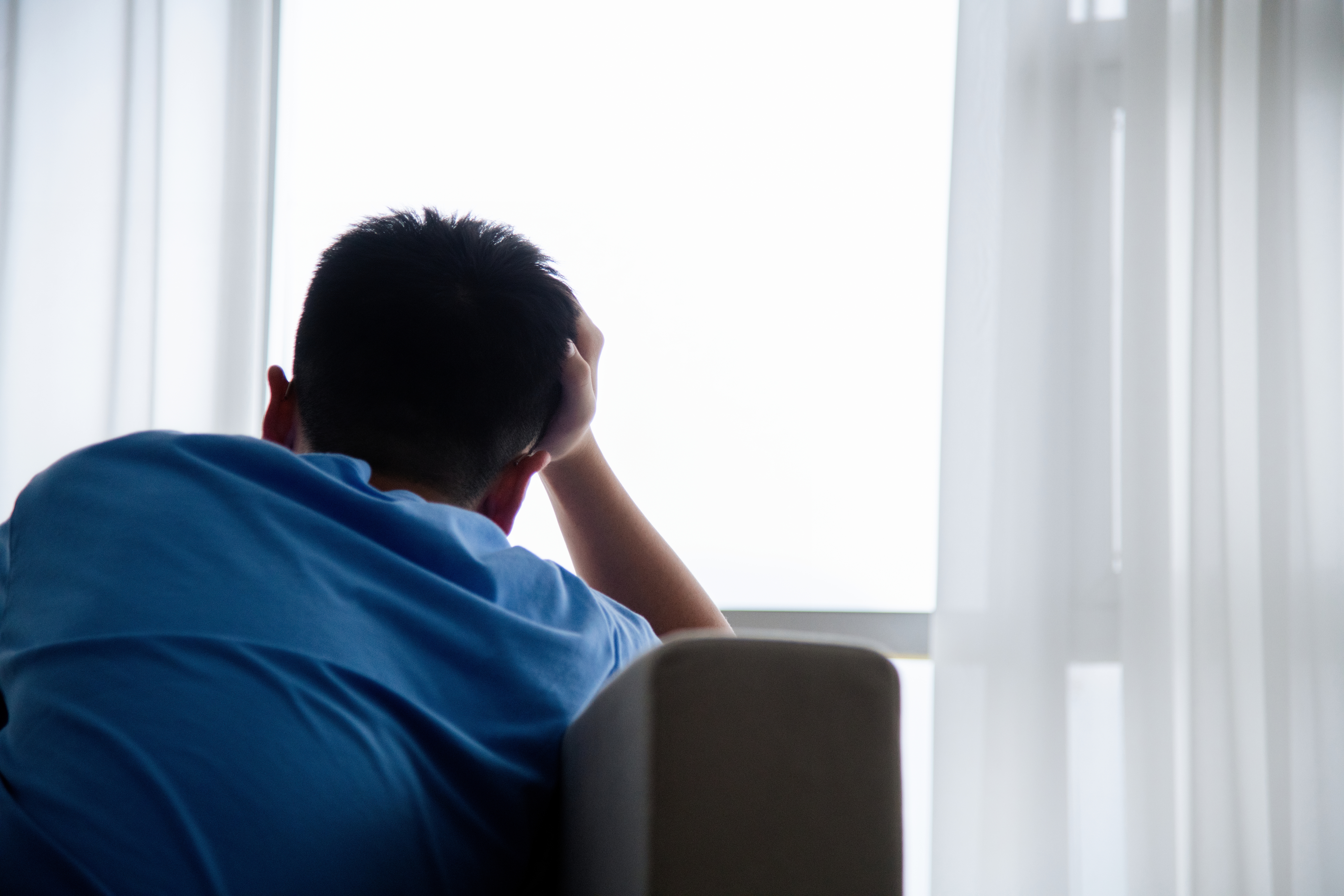 Person sitting indoors, facing a window with head resting on hand, appearing thoughtful or contemplative